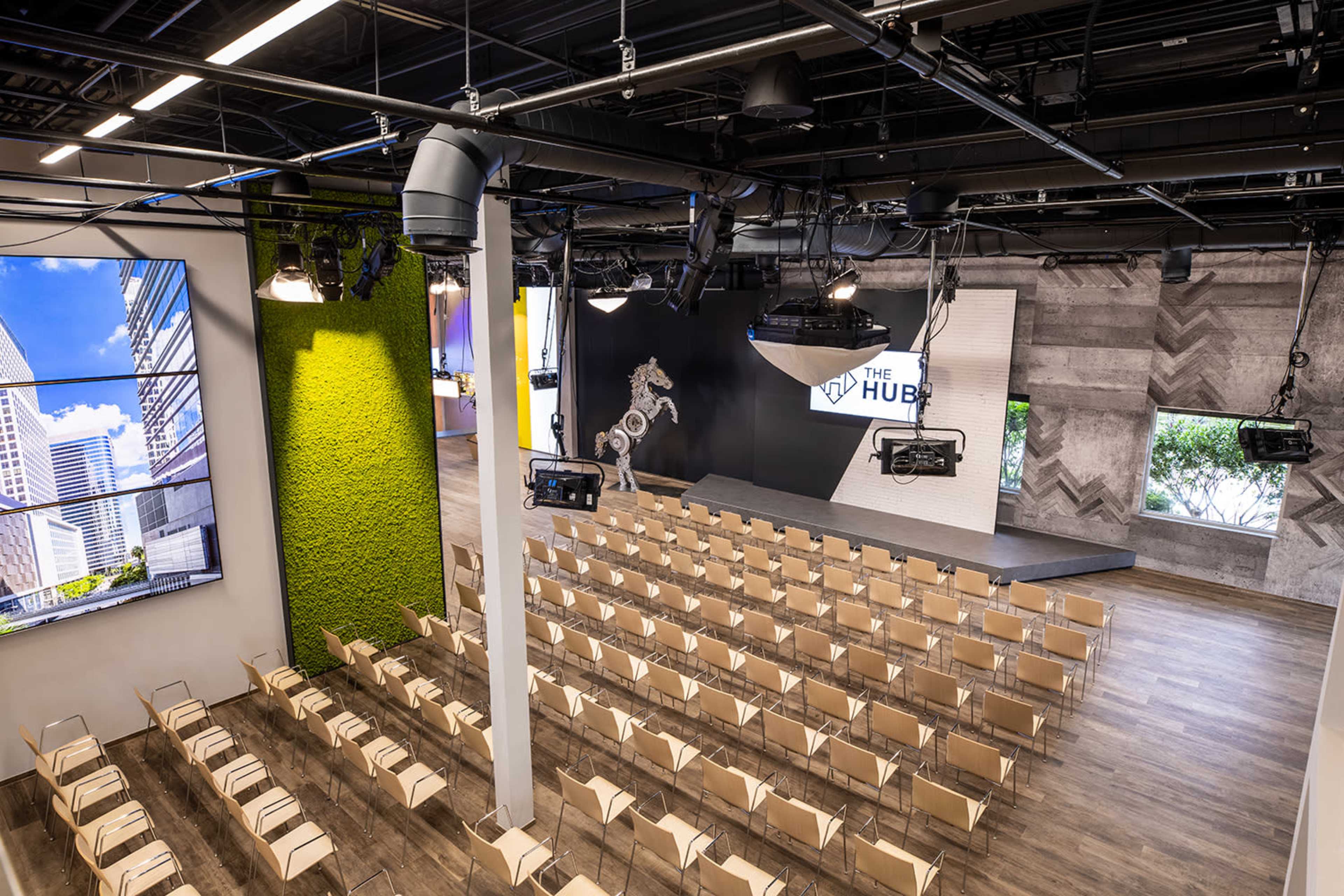 The image shows a modern auditorium space with rows of beige chairs set up in front of a stage featuring a large screen and a decorative sculpture.