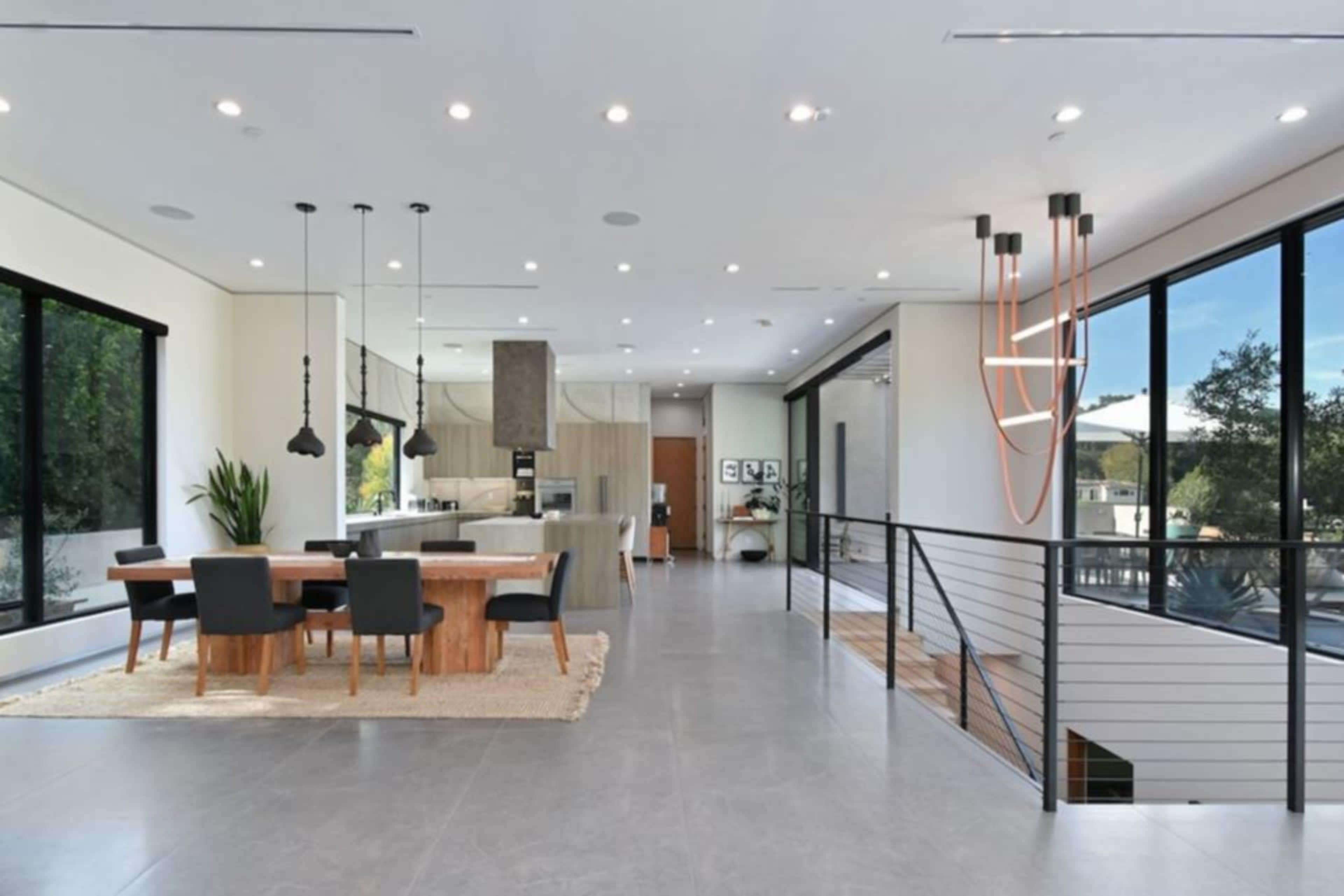 The image shows a spacious, modern open-concept living area featuring a dining table with chairs, a kitchen area, large windows, and minimalist decor.