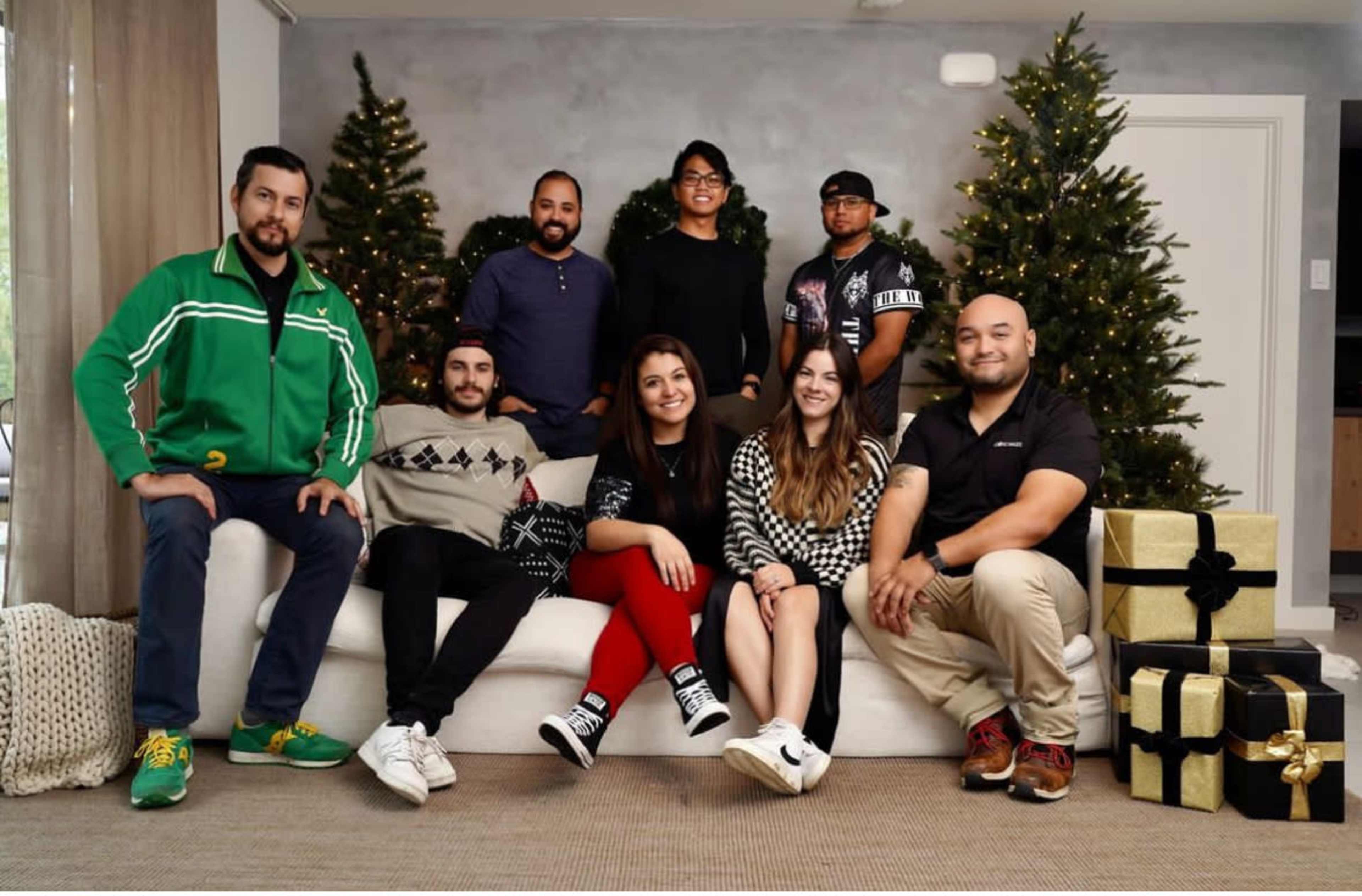 A group of eight people is posing on a couch in front of two decorated Christmas trees and gift boxes.
