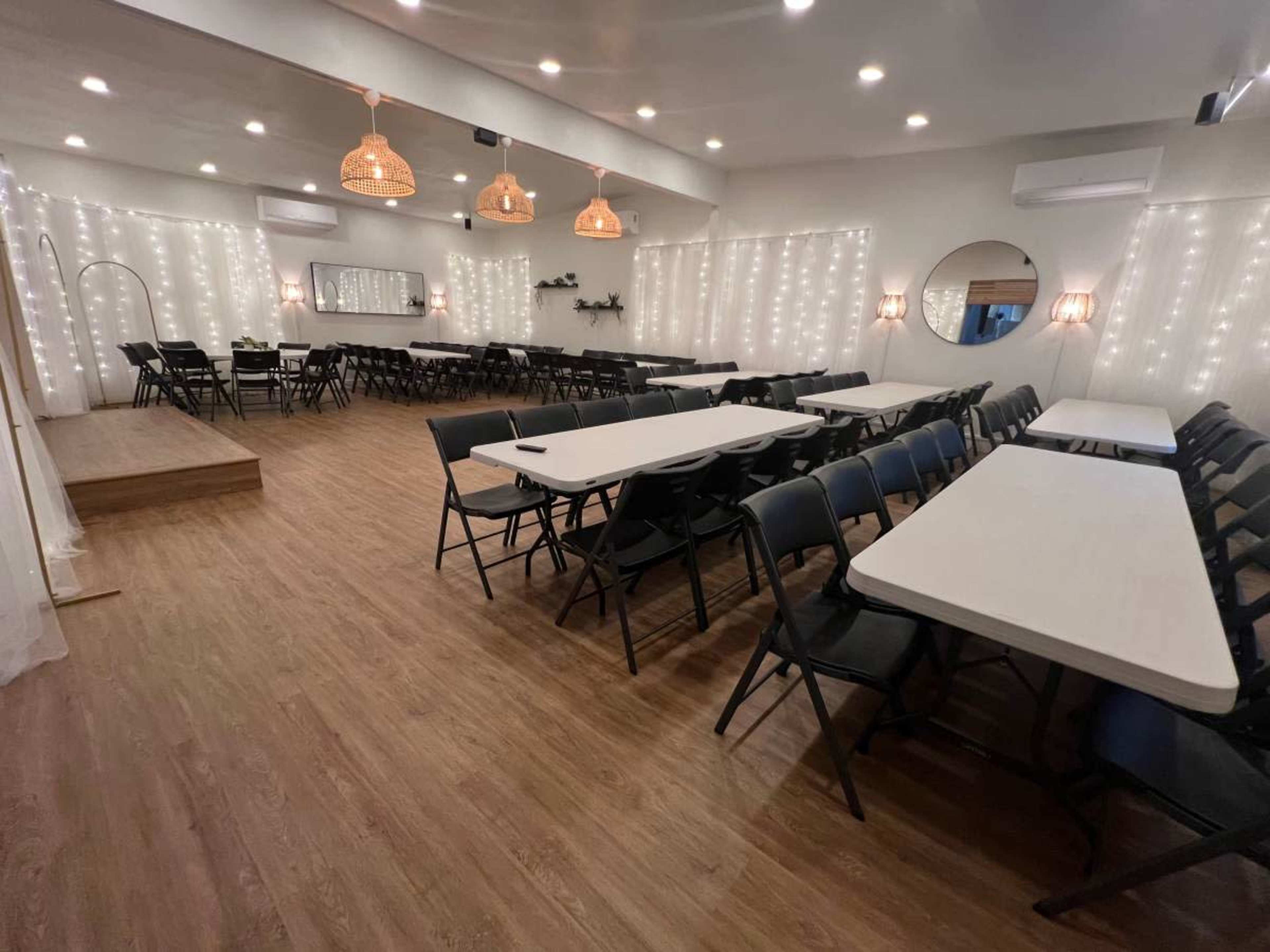 A spacious indoor venue arranged with multiple rectangular tables and black chairs, illuminated by string lights along the walls.