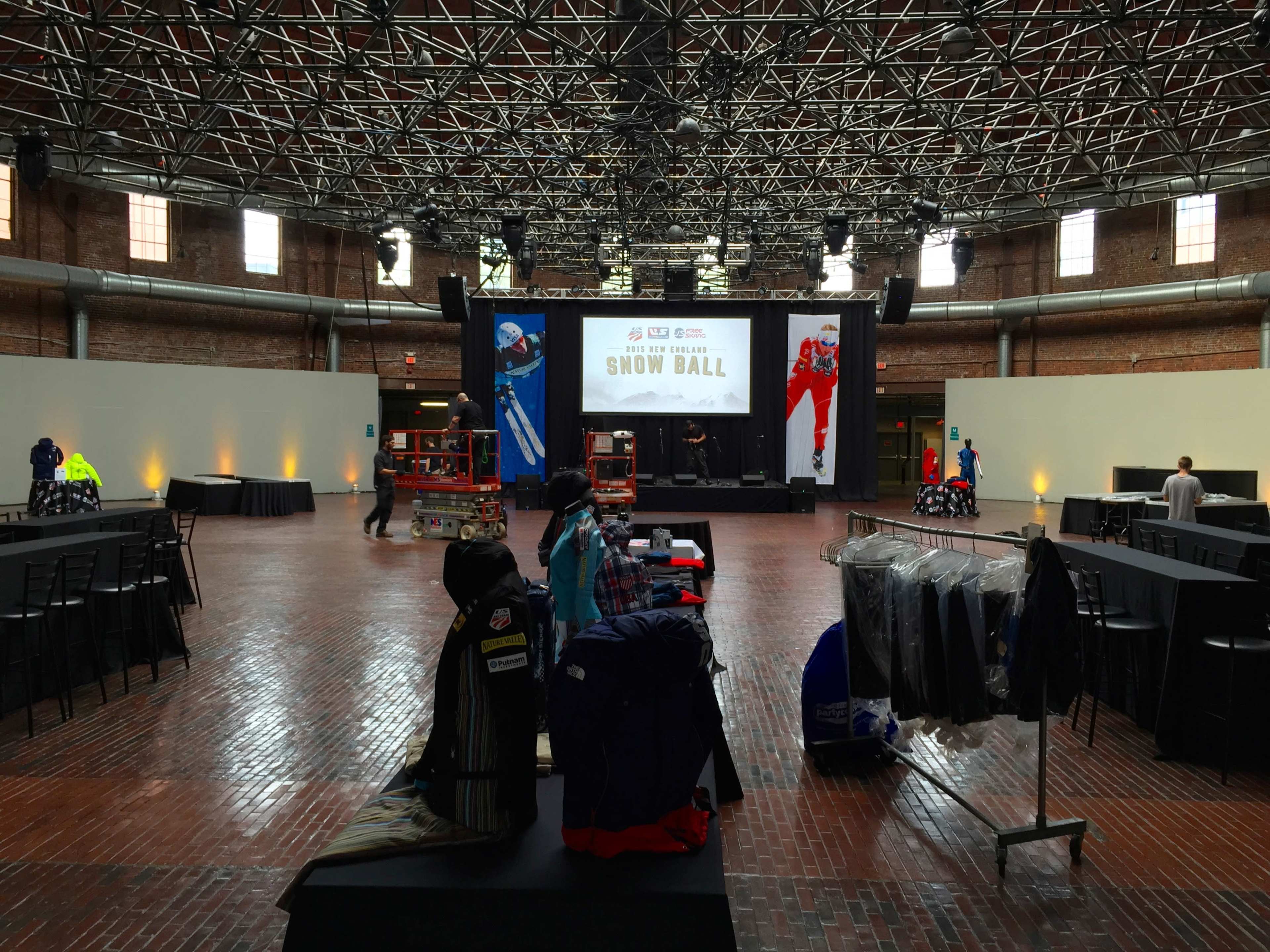 An empty event space with a stage at the front, surrounded by tables and chairs, and various clothing items displayed for an upcoming show.