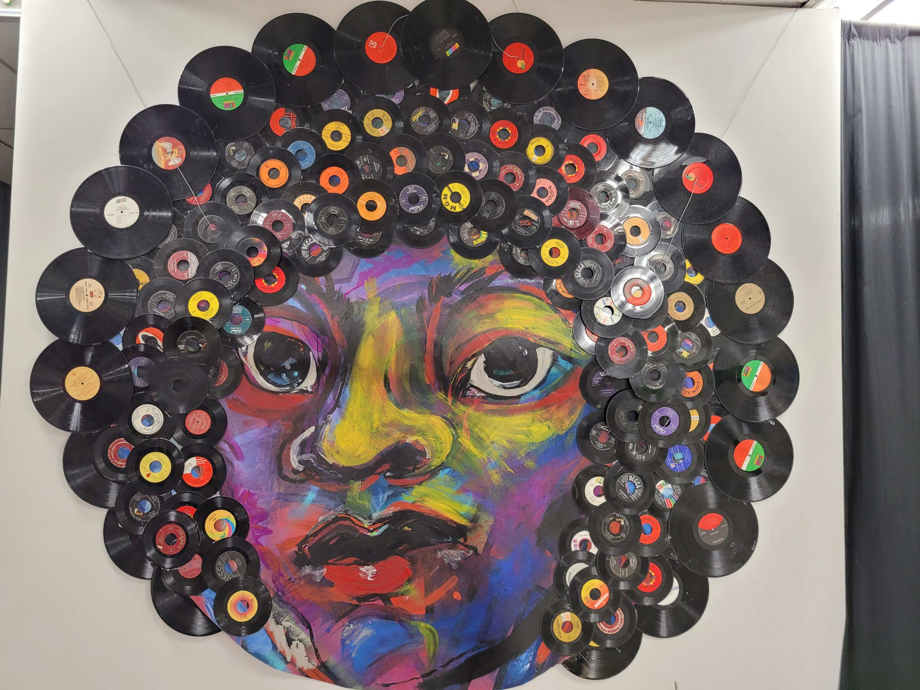 A colorful mural of a face surrounded by an arrangement of vinyl records that form a circular pattern resembling hair.