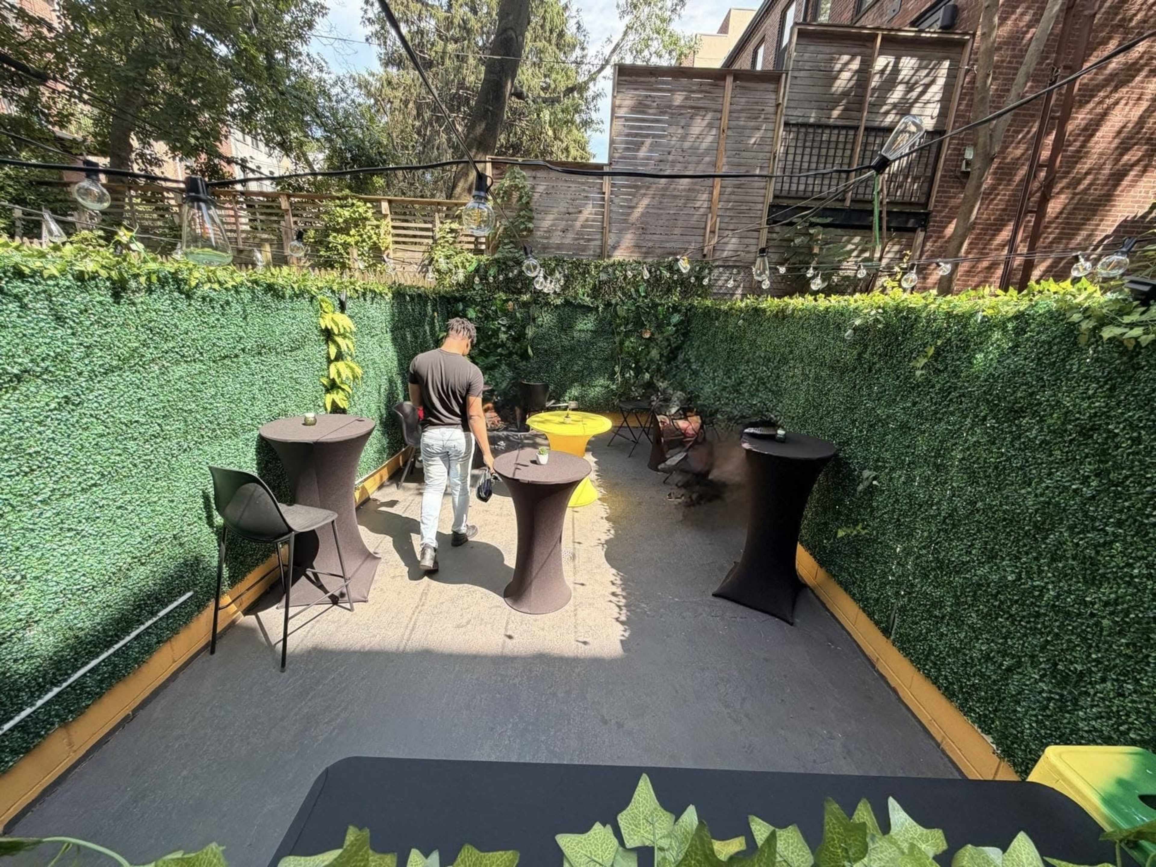 A man sets up tables in a small outdoor space surrounded by tall greenery.