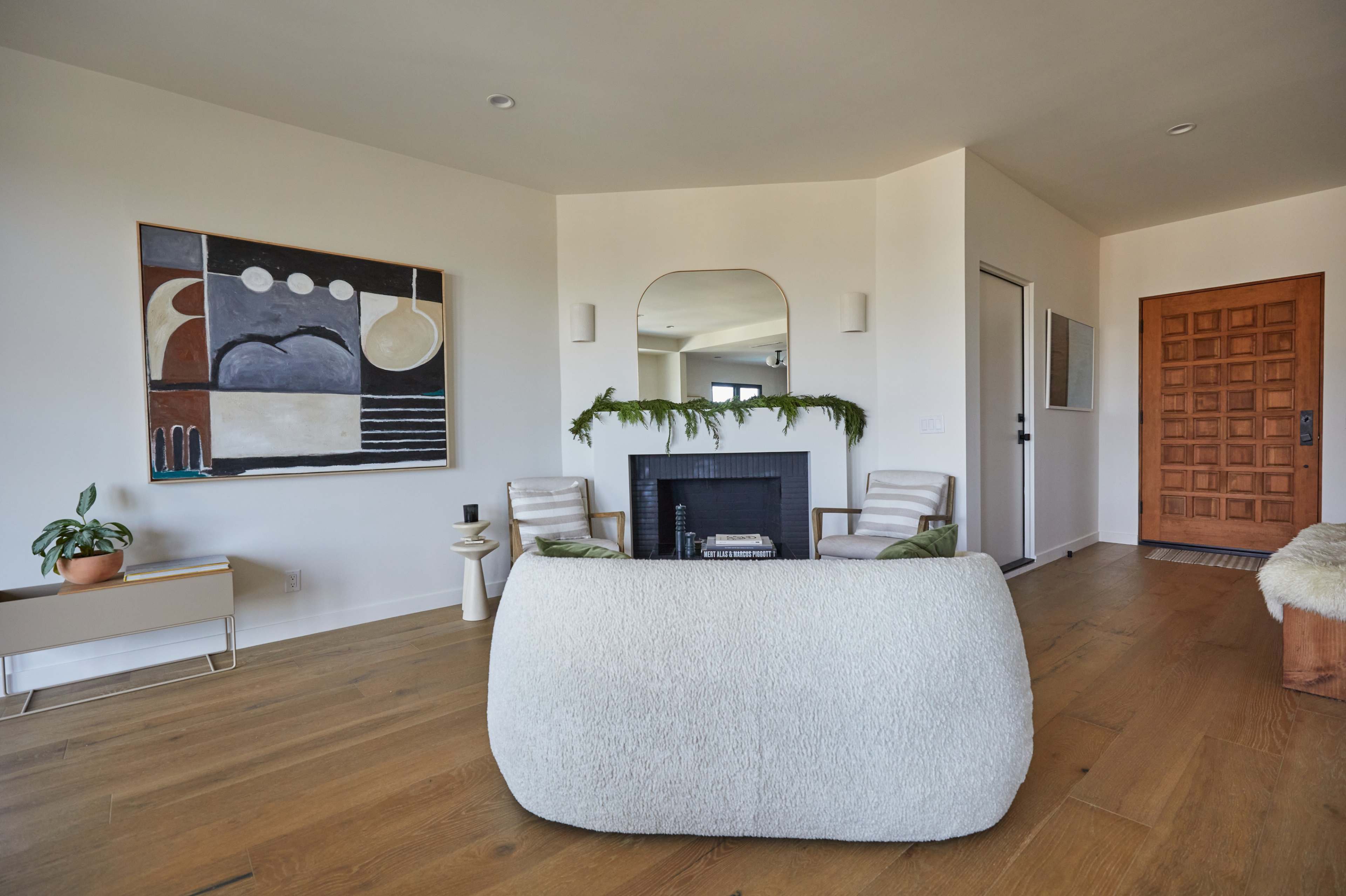 The image shows a modern living room featuring a round, textured sofa, a minimalist fireplace, and a large artwork on the wall, along with a wooden front door.