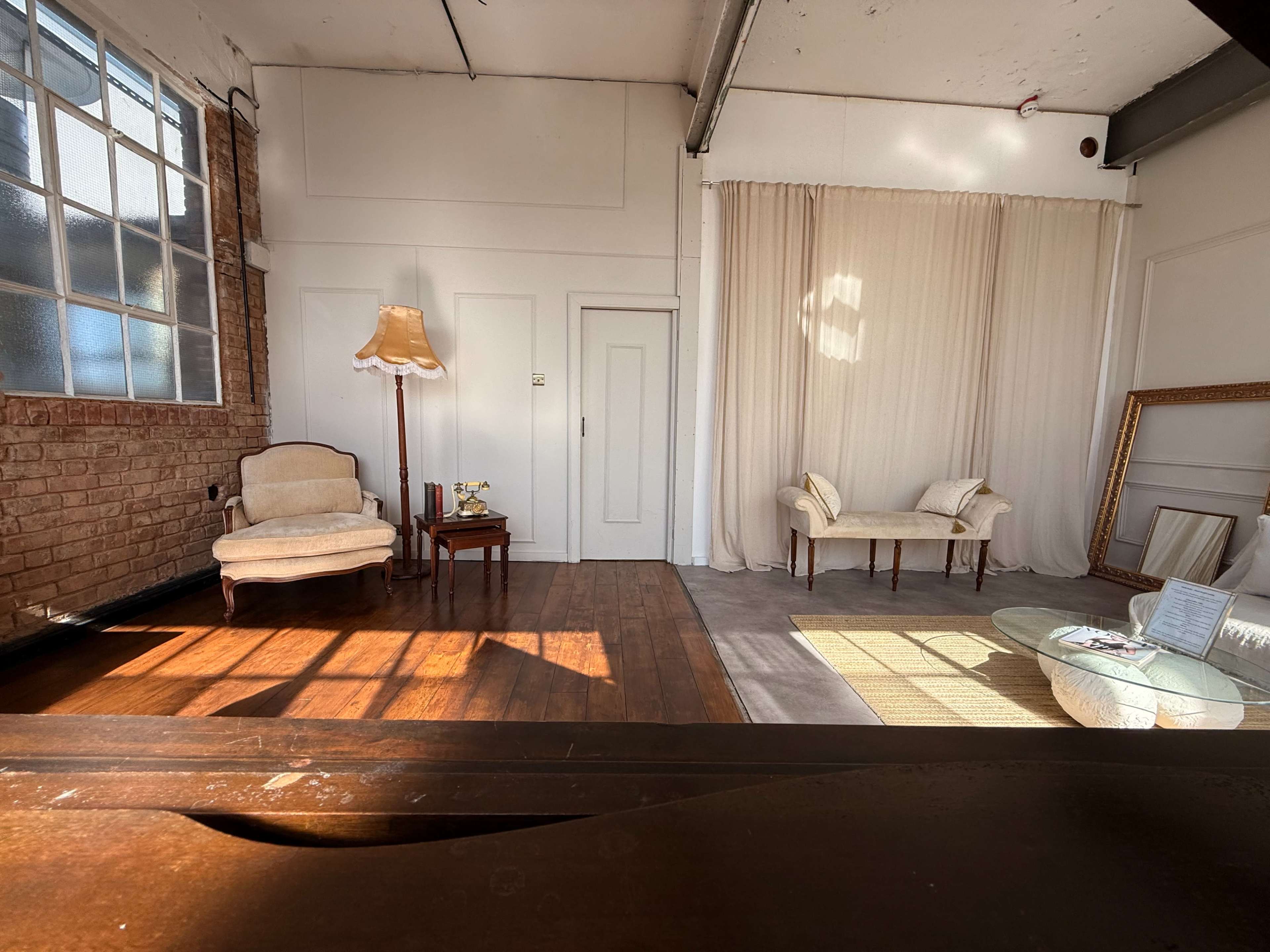 The image shows a spacious interior with wooden flooring, featuring a vintage armchair, a small table with a lamp, and a light-colored curtain draped against the back wall.