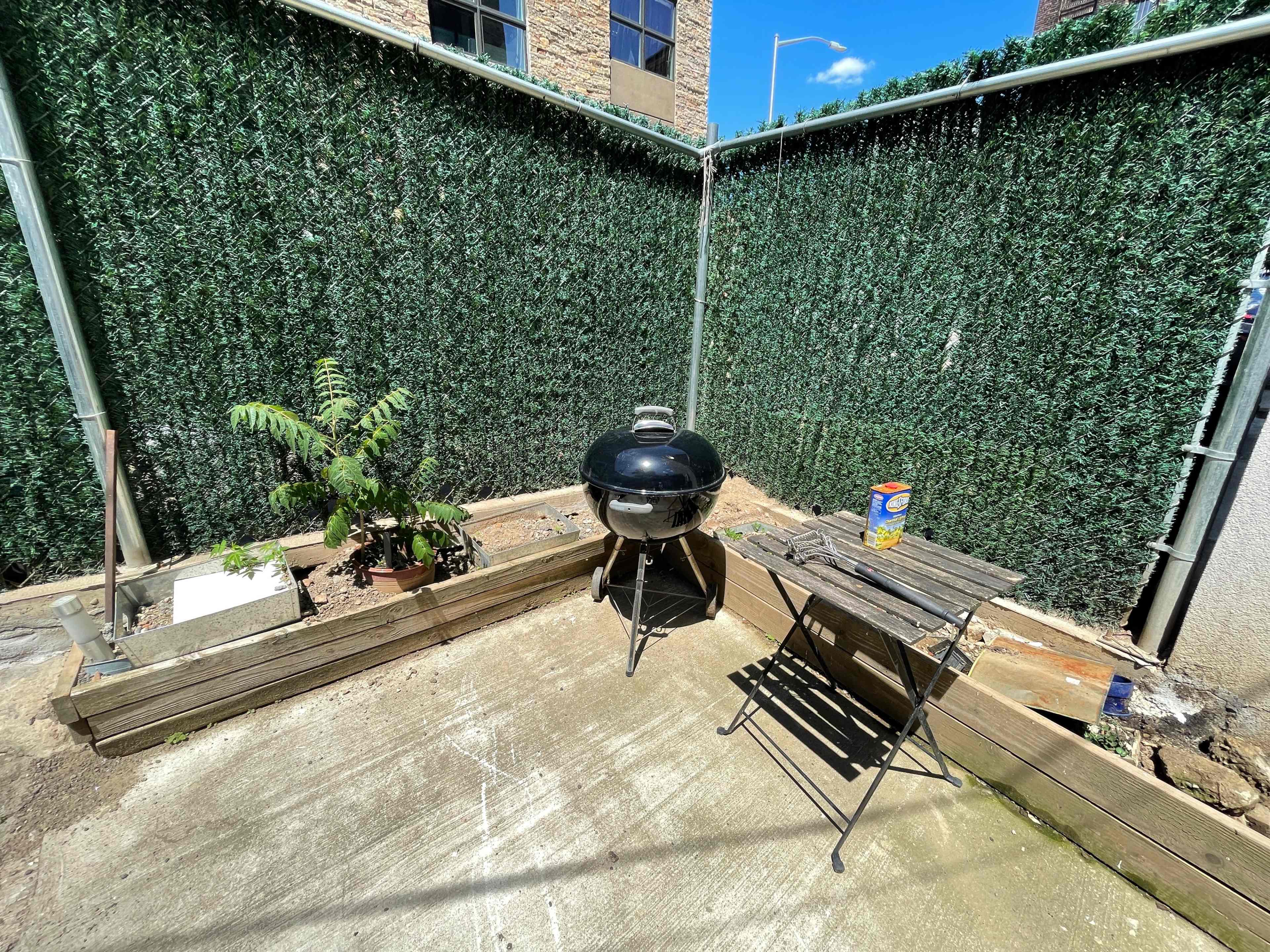 A charcoal grill sits on a small concrete patio surrounded by green privacy fencing, with a potted plant and a folding table nearby.