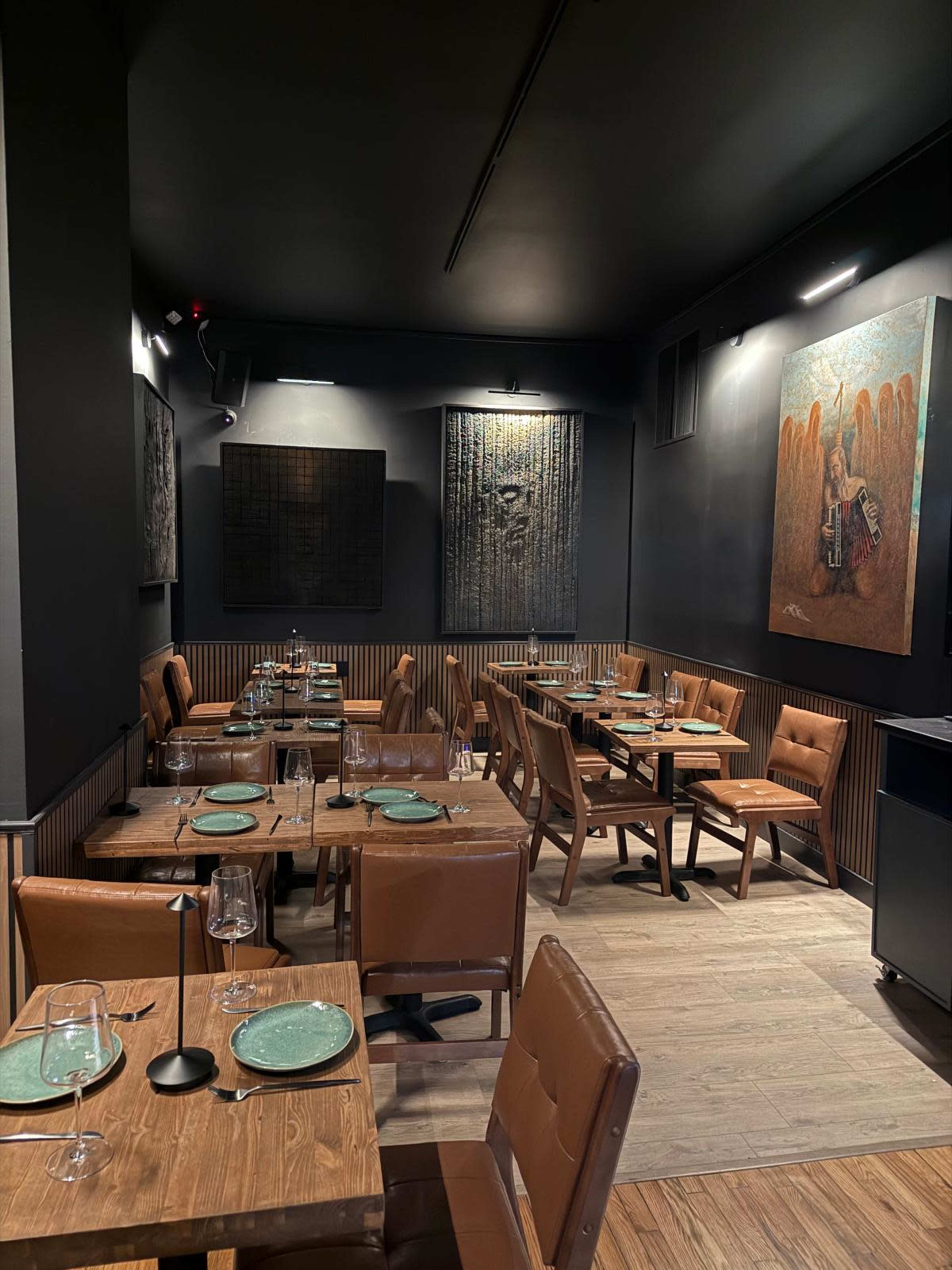 The image shows a dimly lit restaurant with wooden tables, leather chairs, and large artworks displayed on the dark walls.