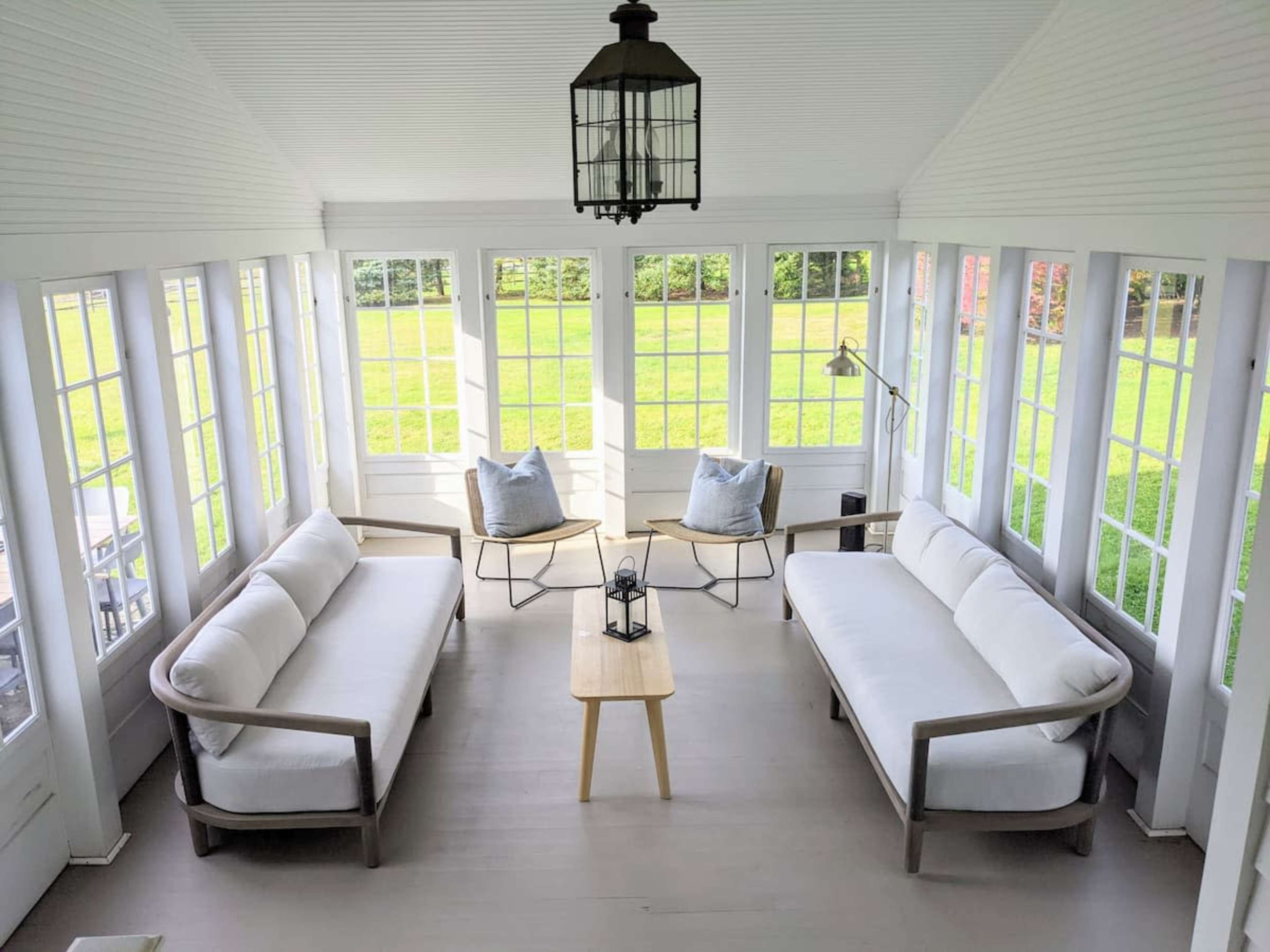 The image shows a bright sunroom with large windows, featuring two white sofas, a small wooden coffee table, and a floor lamp, overlooking a grassy yard.