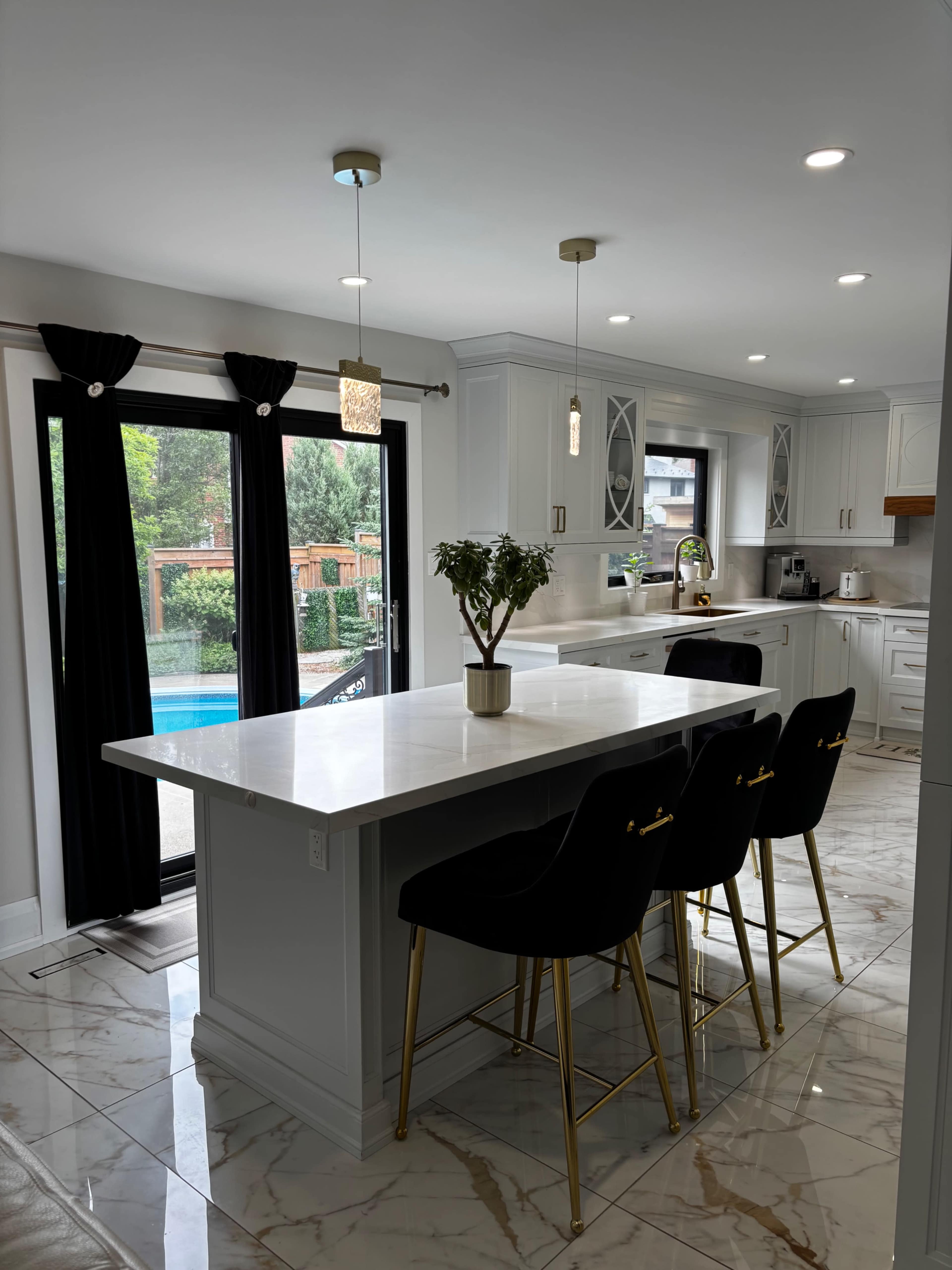 The image shows a modern kitchen featuring a marble island with black bar stools, white cabinetry, and large windows overlooking a pool area.