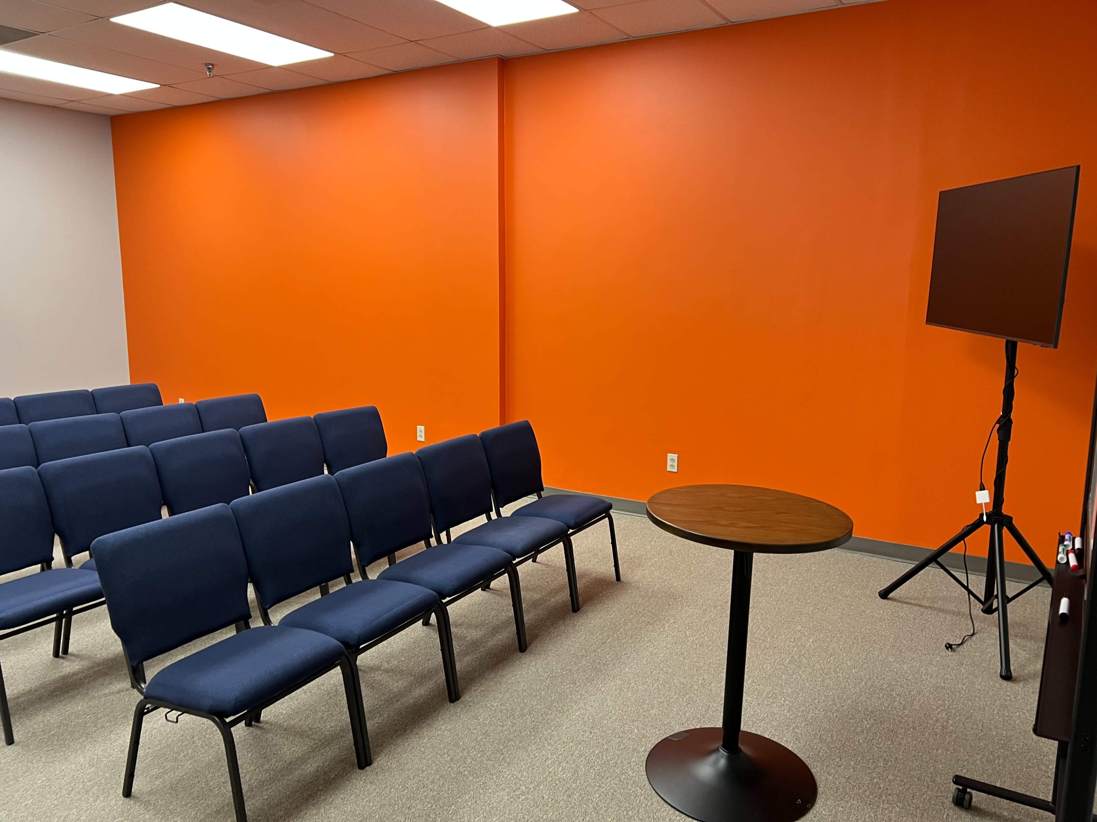 The image shows a small meeting room with blue chairs arranged in rows facing a circular table and a mounted screen against a bright orange wall.