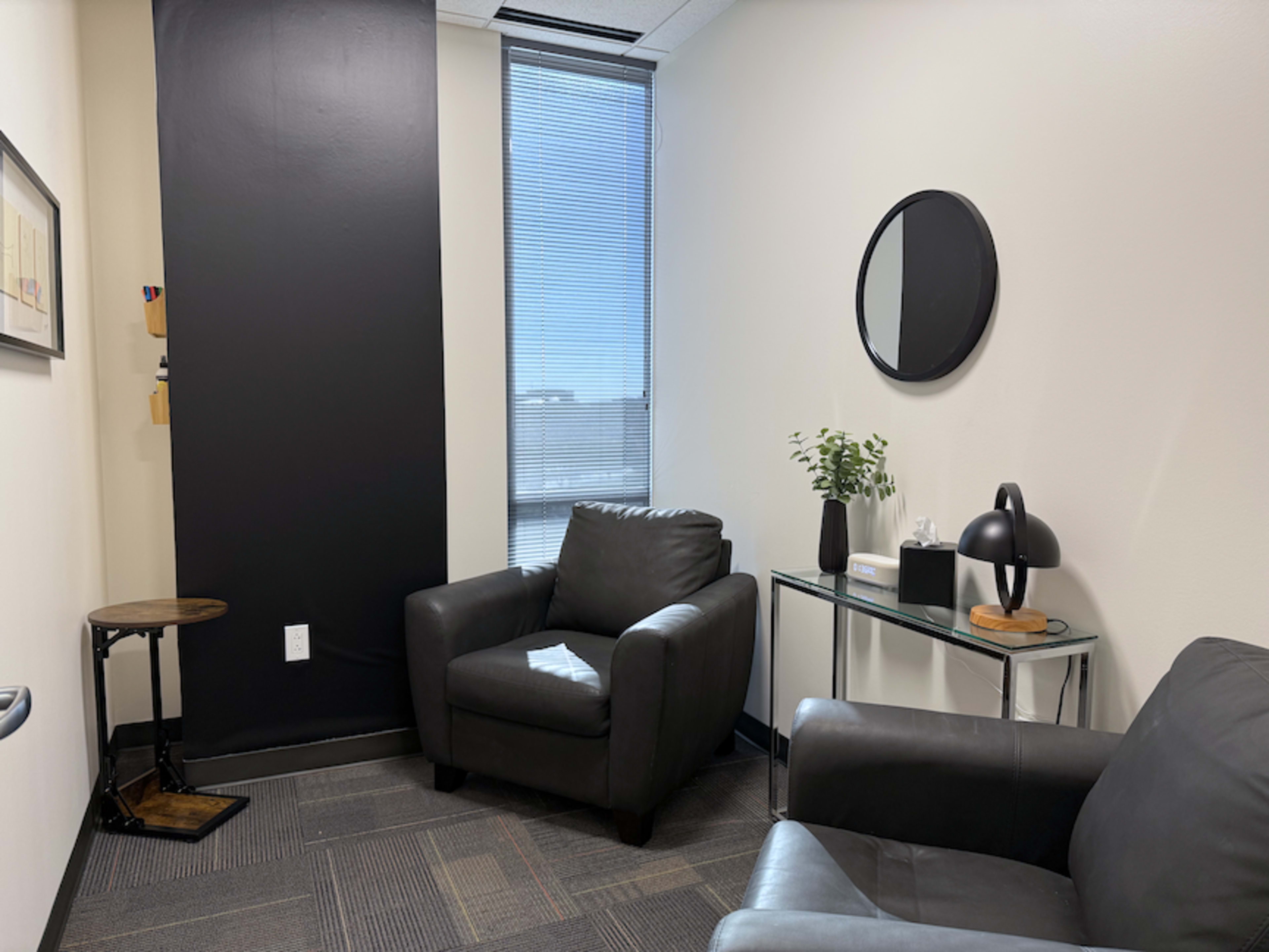The image shows a small office space with two leather armchairs, a glass side table, a lamp, and a window with a view of a clear sky.