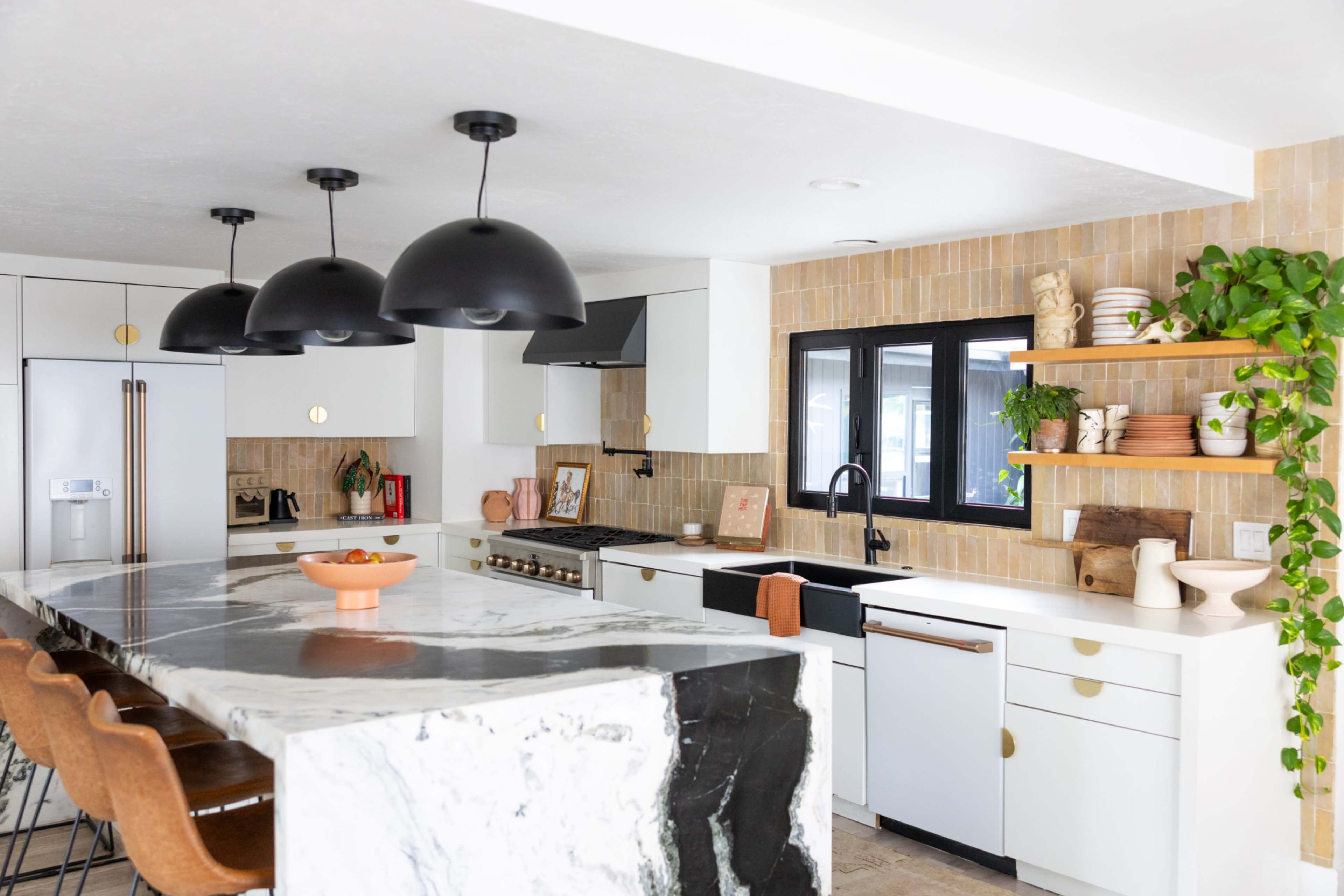 The kitchen features a large island with a marble countertop, black pendant lights, and modern cabinetry, accented by warm wood shelves and greenery.