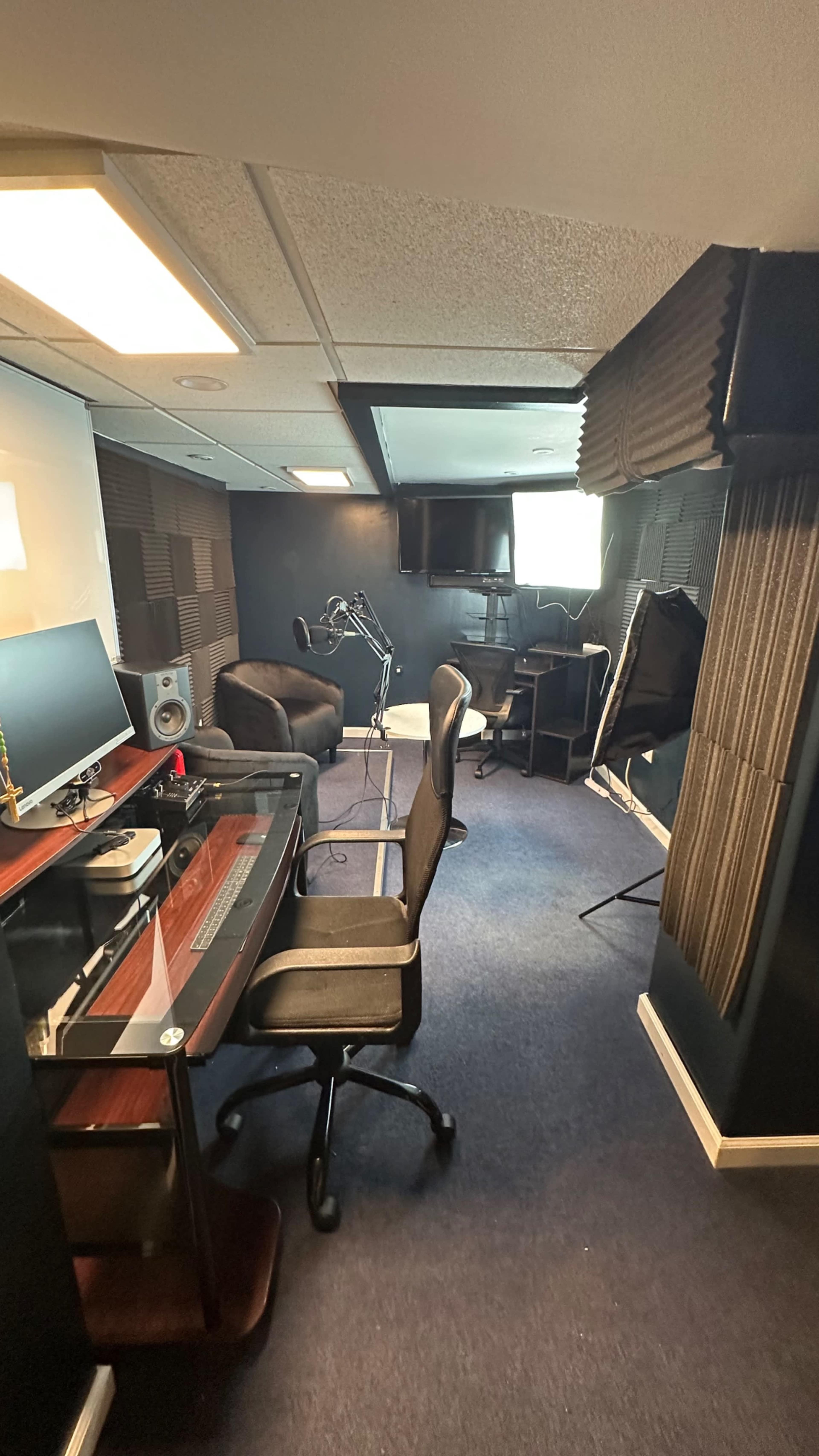 A small recording studio featuring a desk with a computer, two chairs, a microphone setup, and soundproofing panels on the walls.