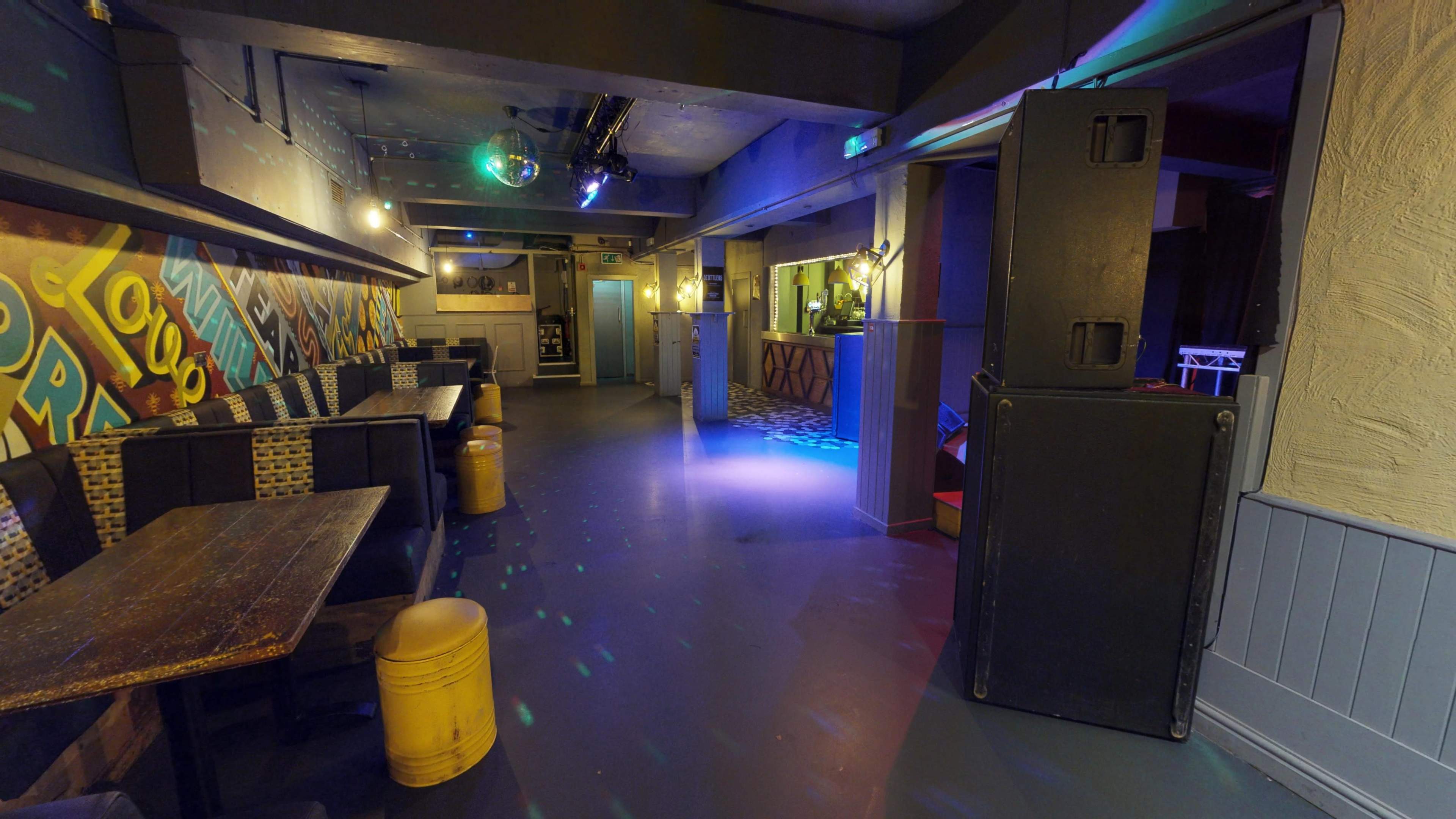 A dimly lit bar or club interior featuring a dance area with a disco ball, tables along the walls, and a bar counter at the far end.