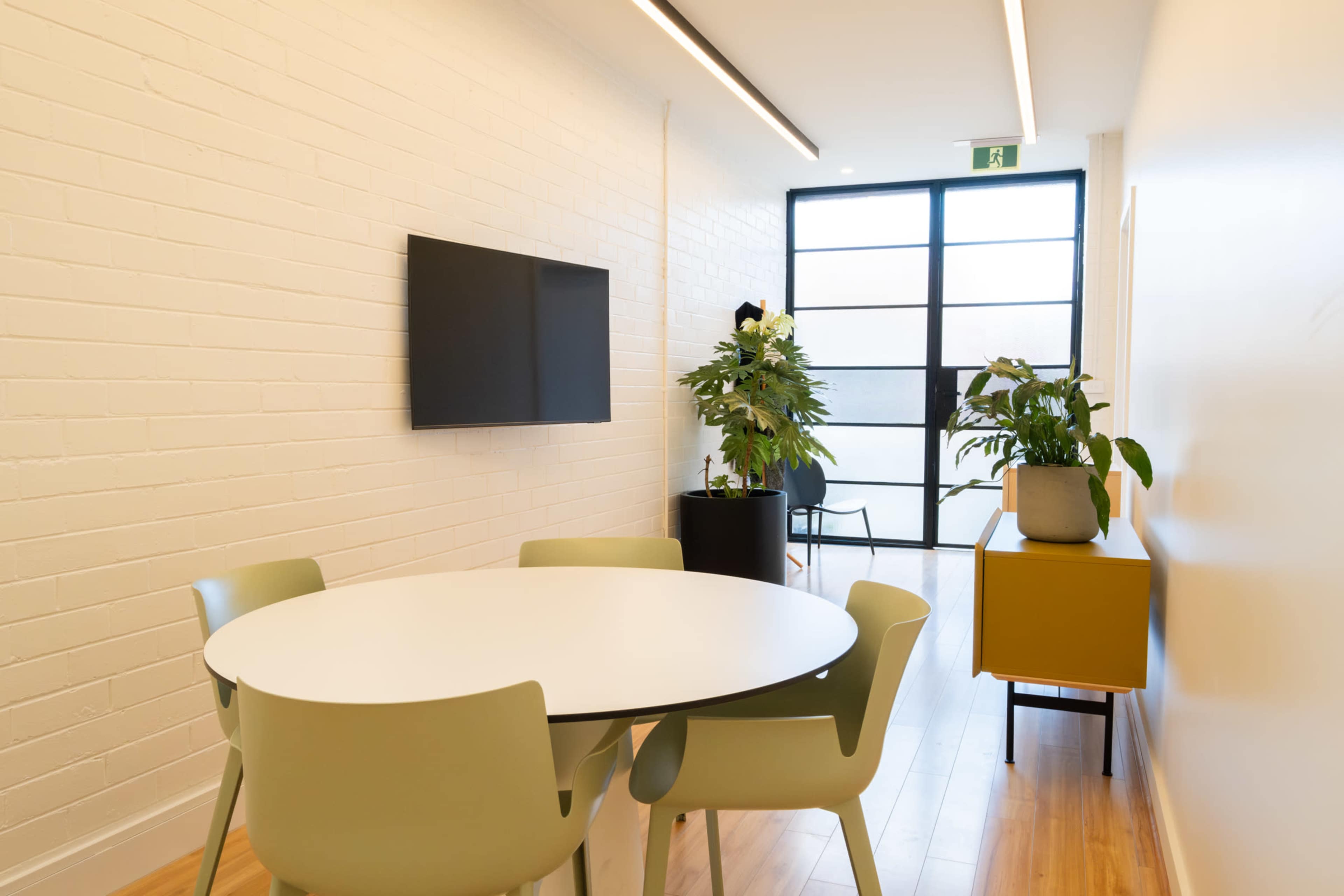 A modern conference room features a round white table surrounded by light-colored chairs, with a wall-mounted TV and potted plants visible.