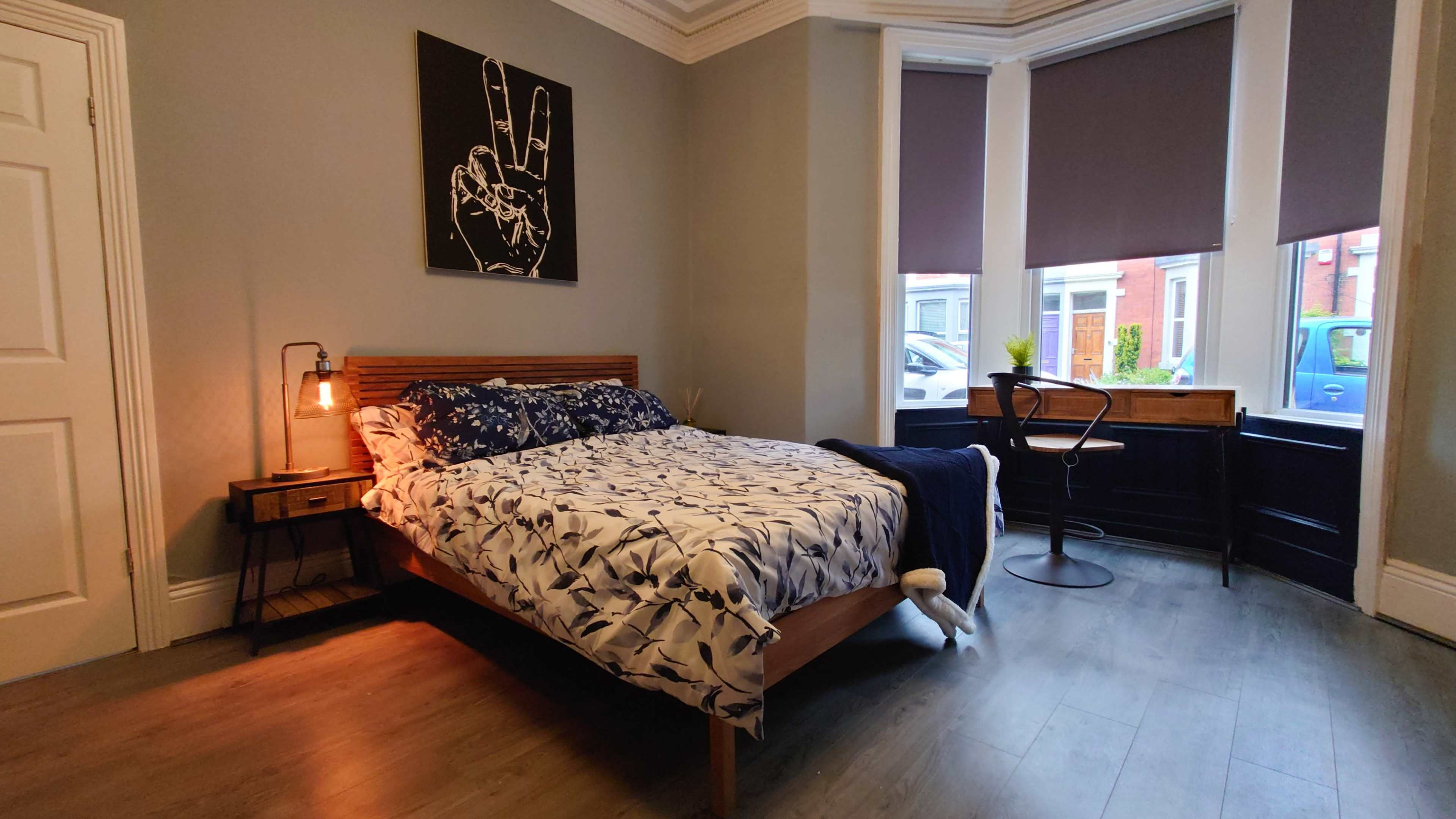 A cozy bedroom features a wooden bed with patterned bedding, a bedside table with a lamp, and a desk by the window.