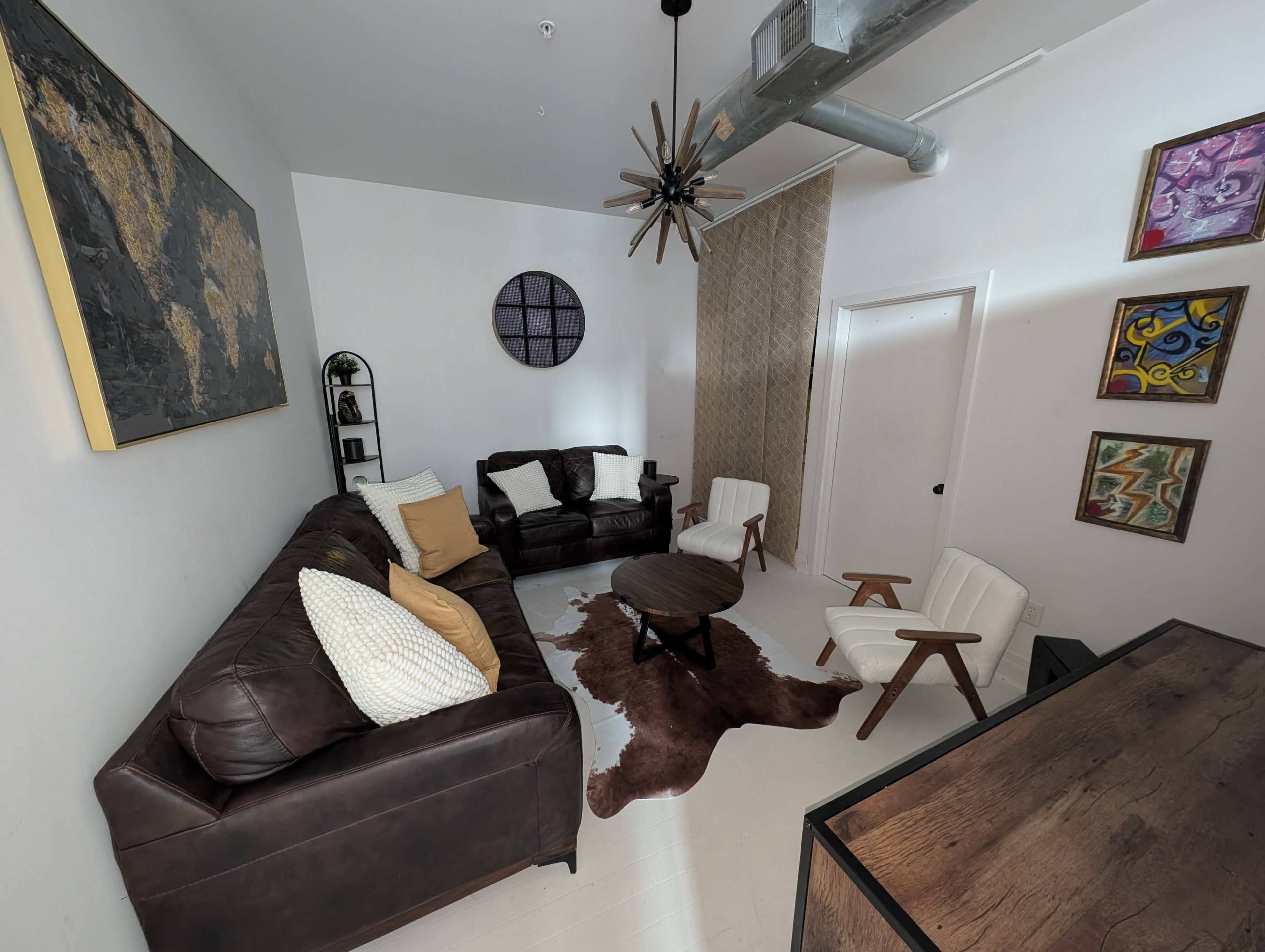The image shows a modern living room with brown leather sofas, a round wooden coffee table, and decorative wall art.