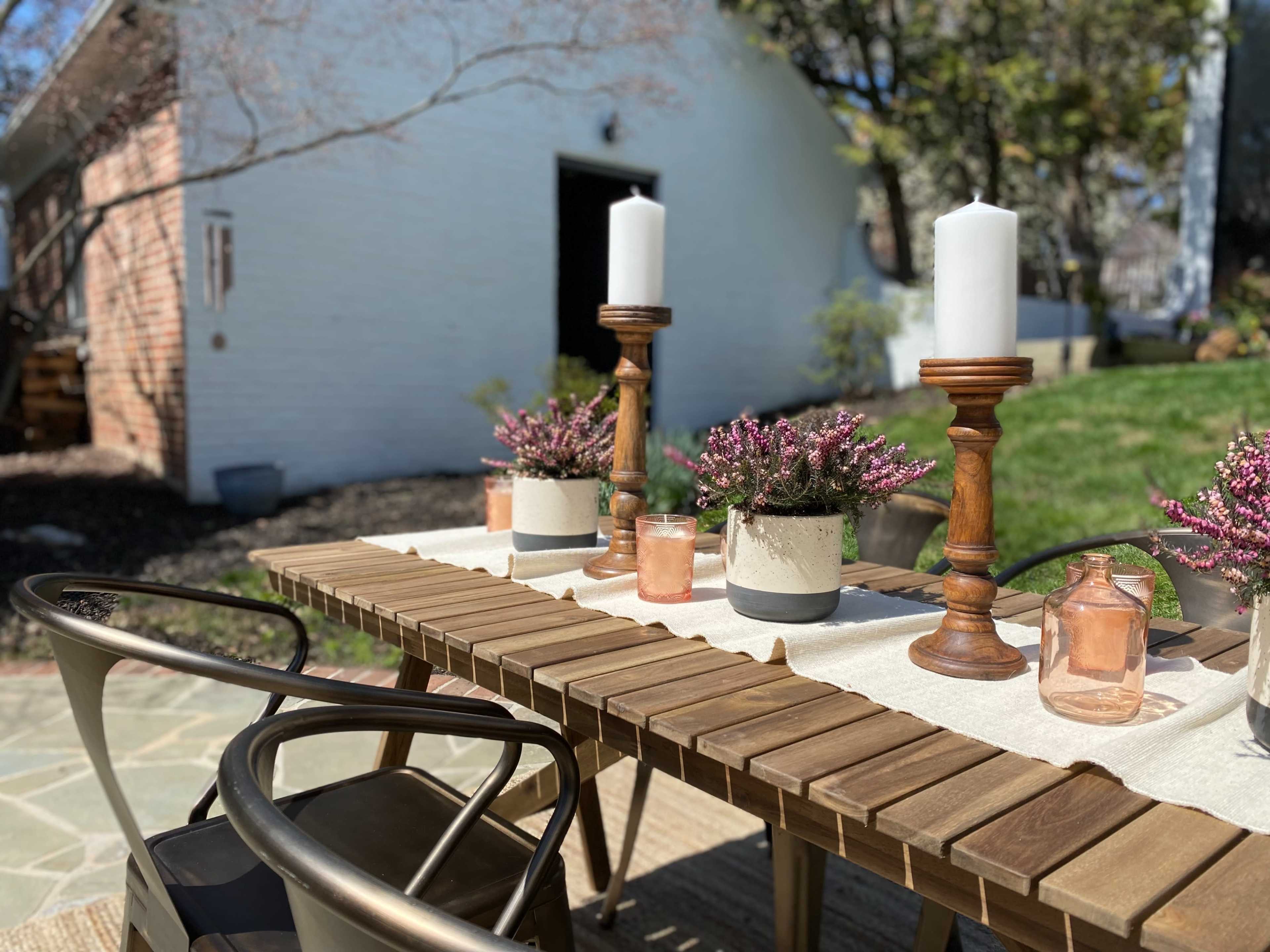 A wooden outdoor dining table is set with candles and potted plants, near a white brick building and garden.