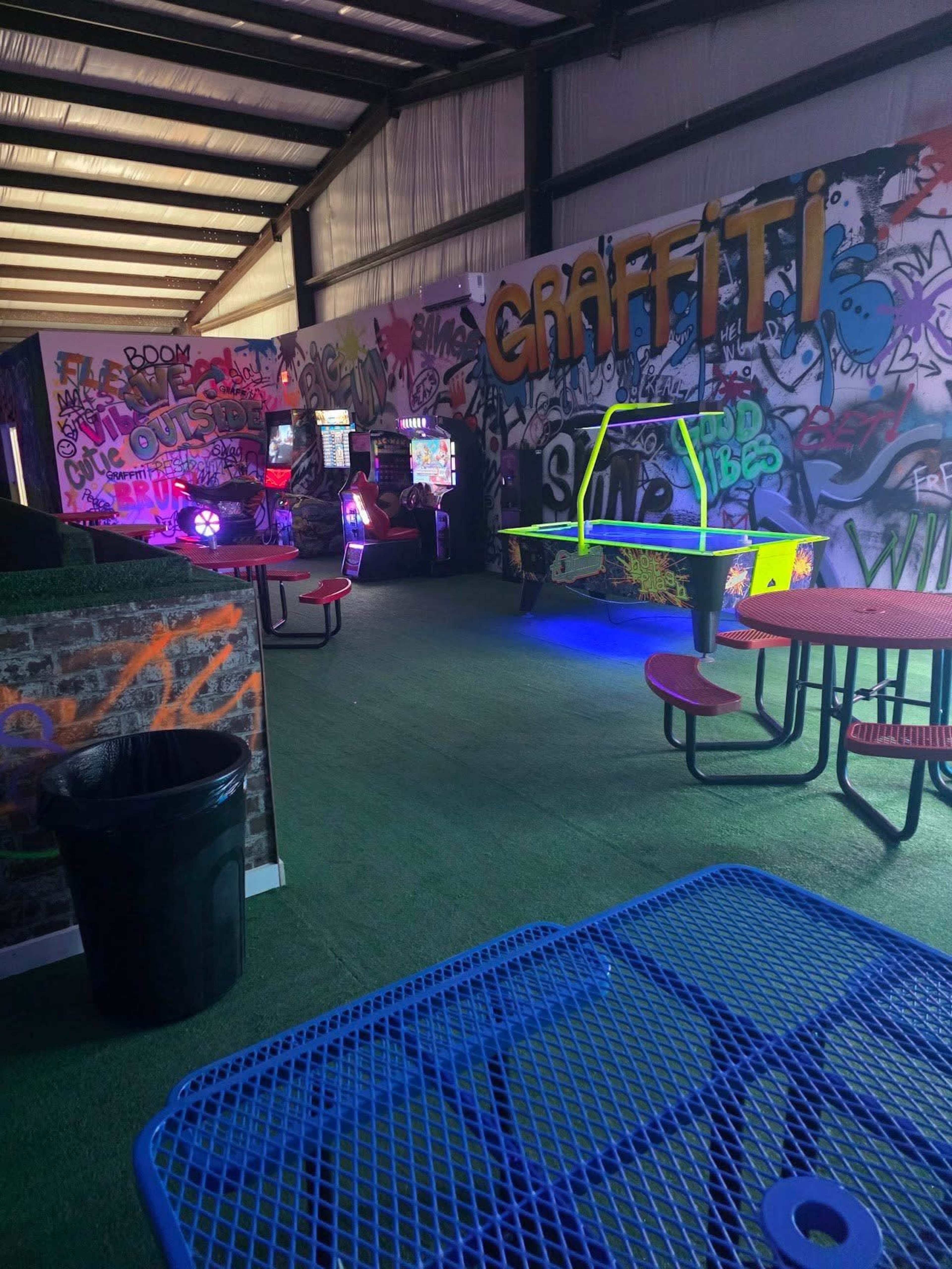 The image shows an indoor space with colorful graffiti on the walls, arcade machines in the background, and tables with chairs arranged on a green carpeted floor.