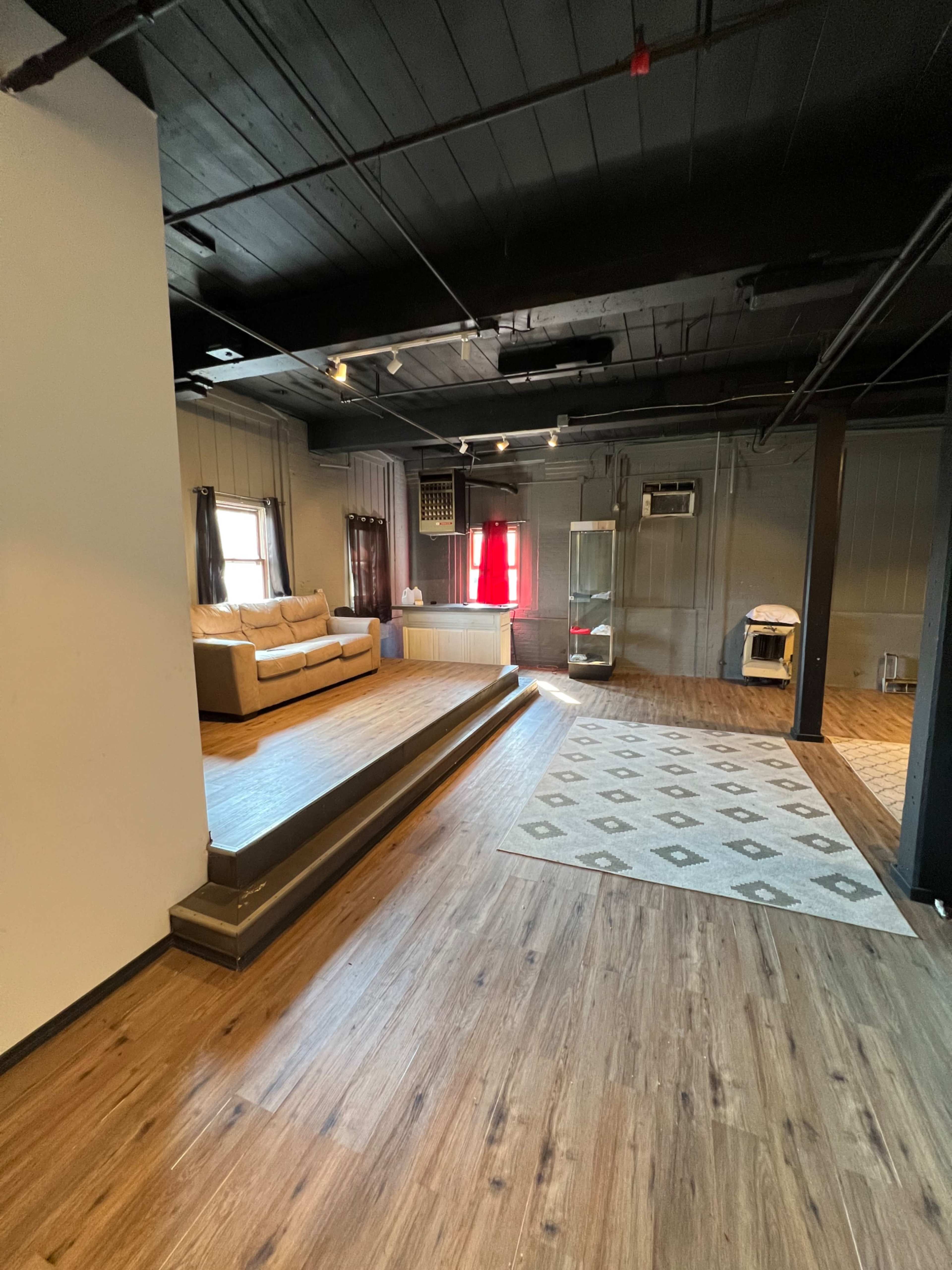 An empty, modern interior space featuring a couch, a small stage area, and a large rug on a wooden floor.