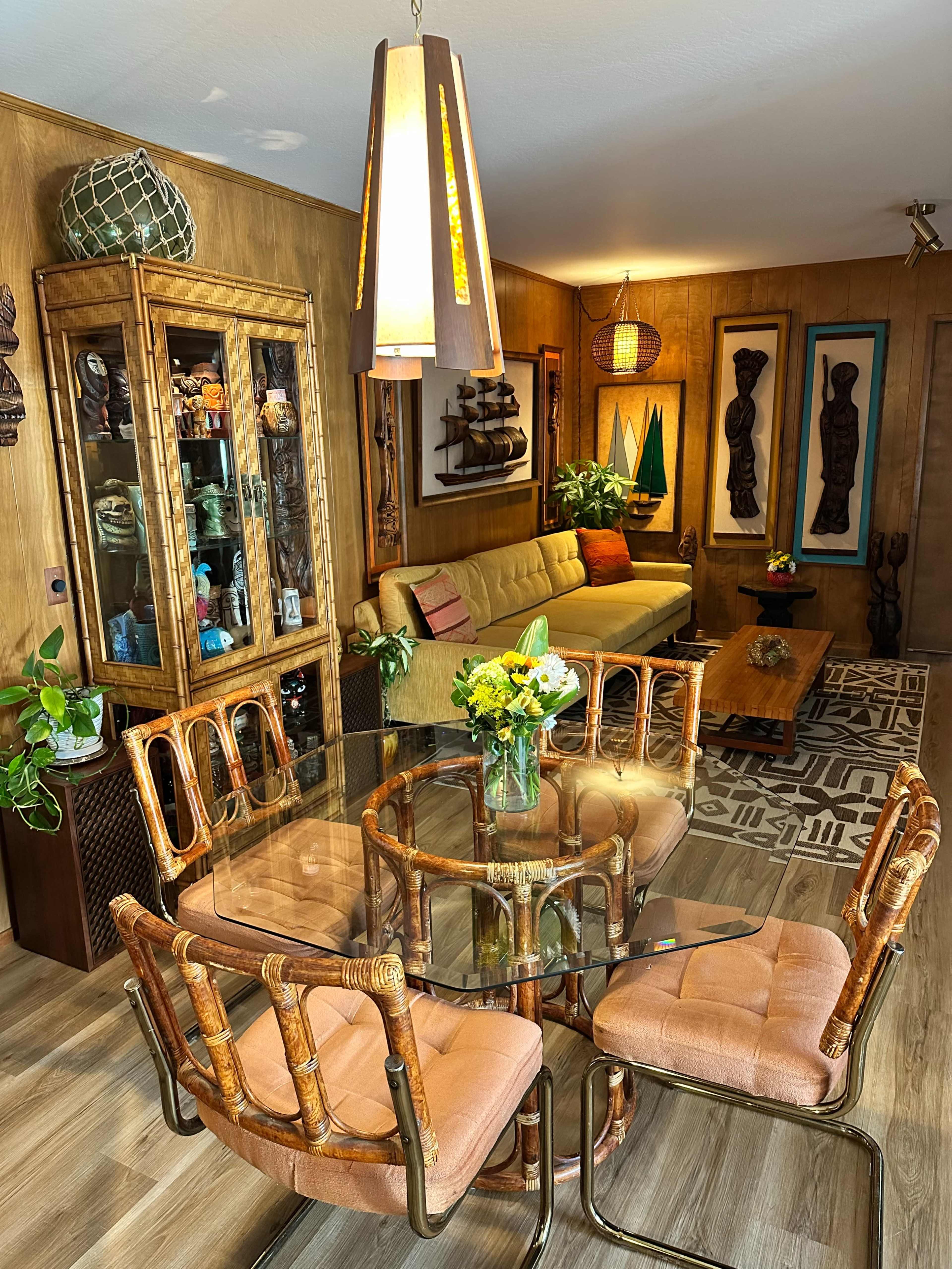 A glass dining table surrounded by upholstered chairs is set in a wood-paneled room featuring additional seating and decorative art pieces.