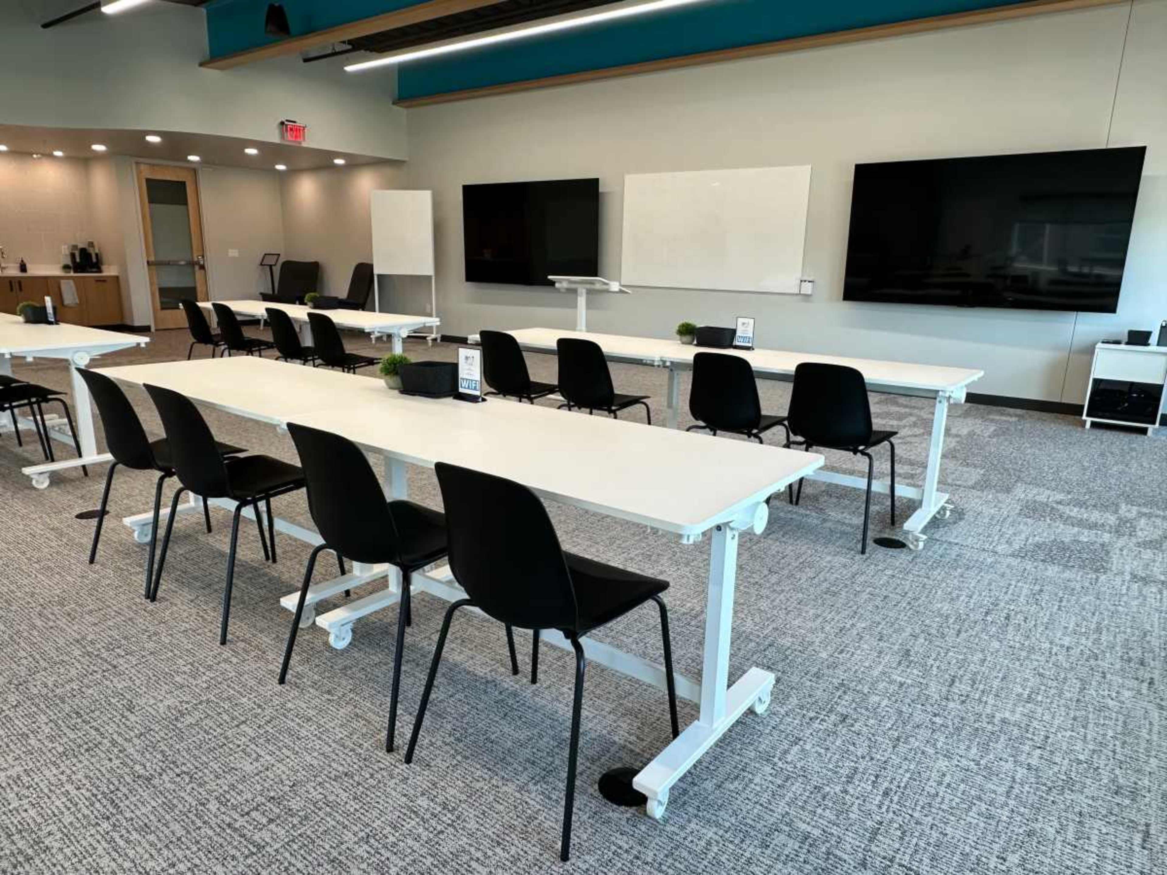The image shows a modern training room with multiple white tables arranged in rows and black chairs, featuring large screens and whiteboards on the walls.