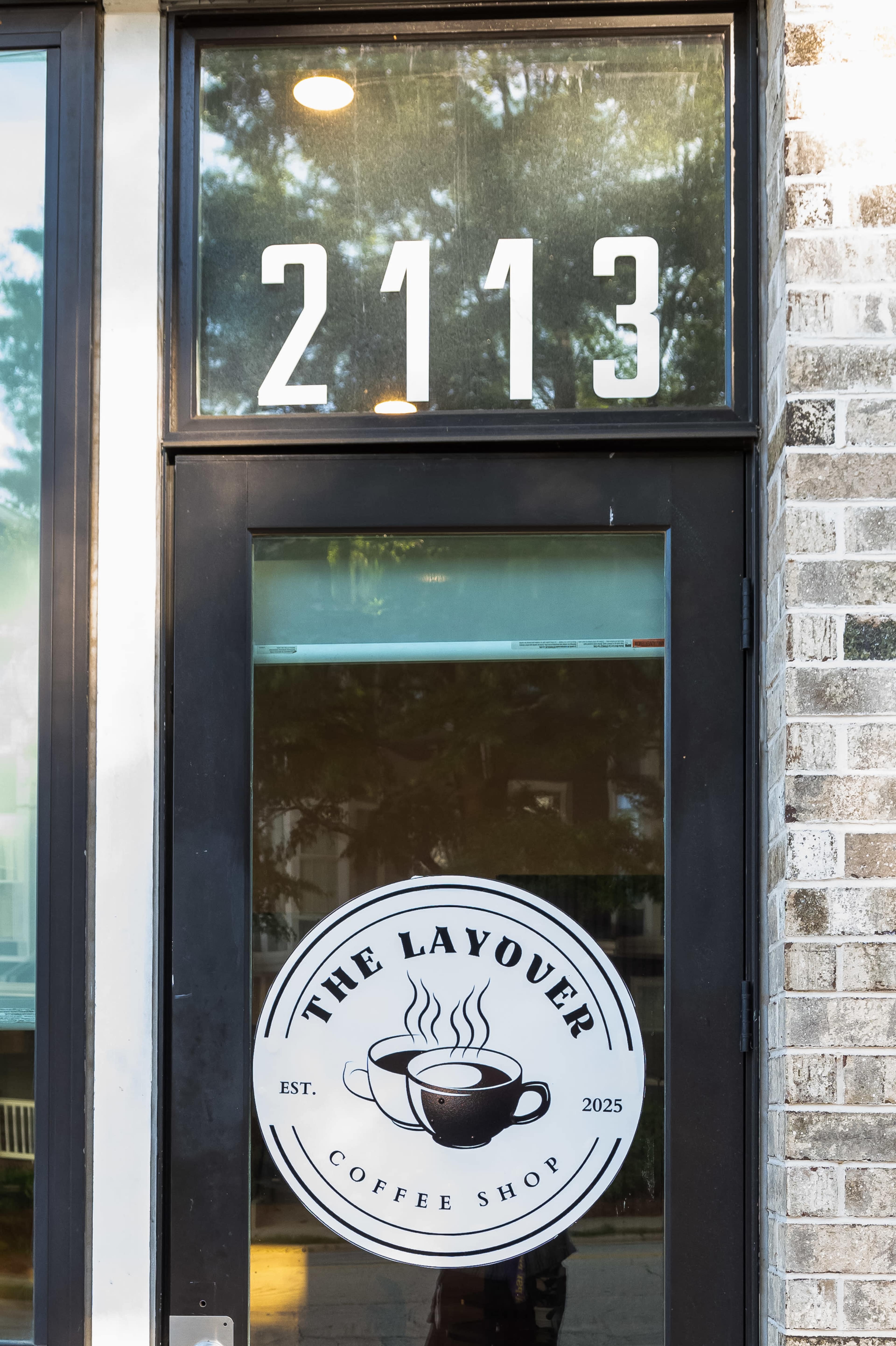 The image shows the entrance of a coffee shop with a window displaying the shop's name, "THE LAYOUEUR," and the establishment year "2025," along with the address "2113."