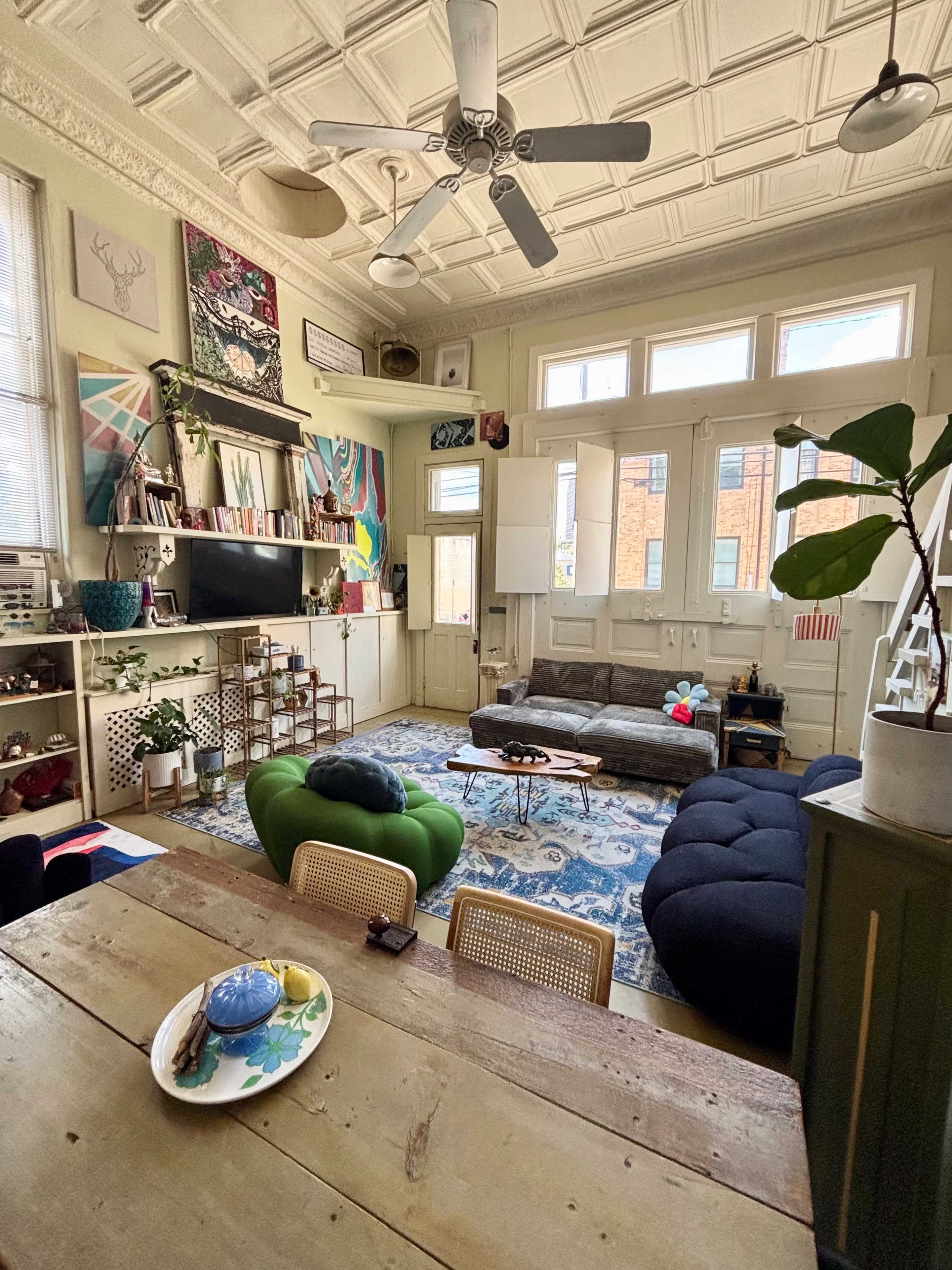 The image shows a brightly lit living room with a mix of modern furniture, colorful decor, and a large patterned rug.