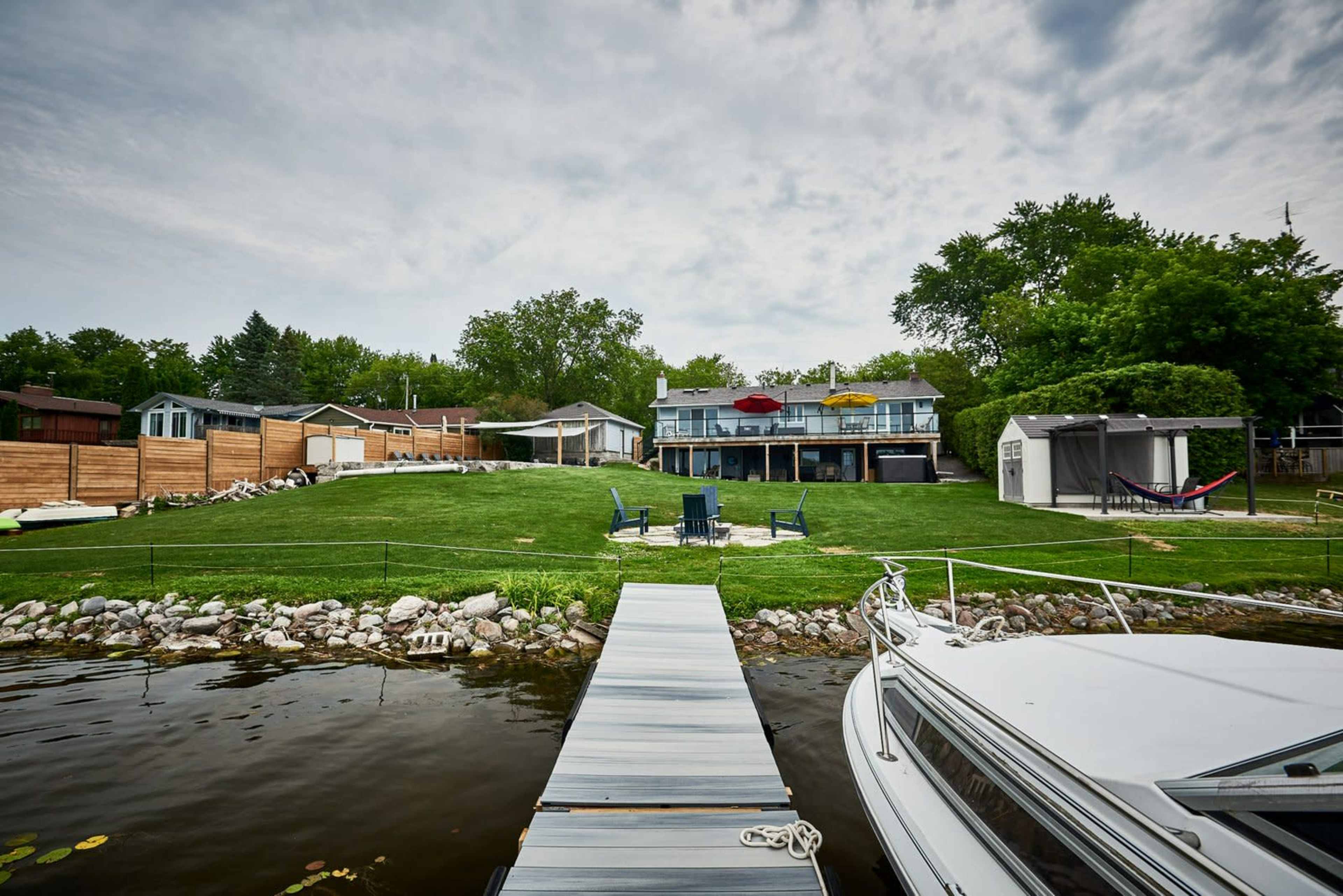 A lakeside property with a wooden dock, a boat moored nearby, and a lawn leading up to a two-story house with outdoor seating and trees in the background.
