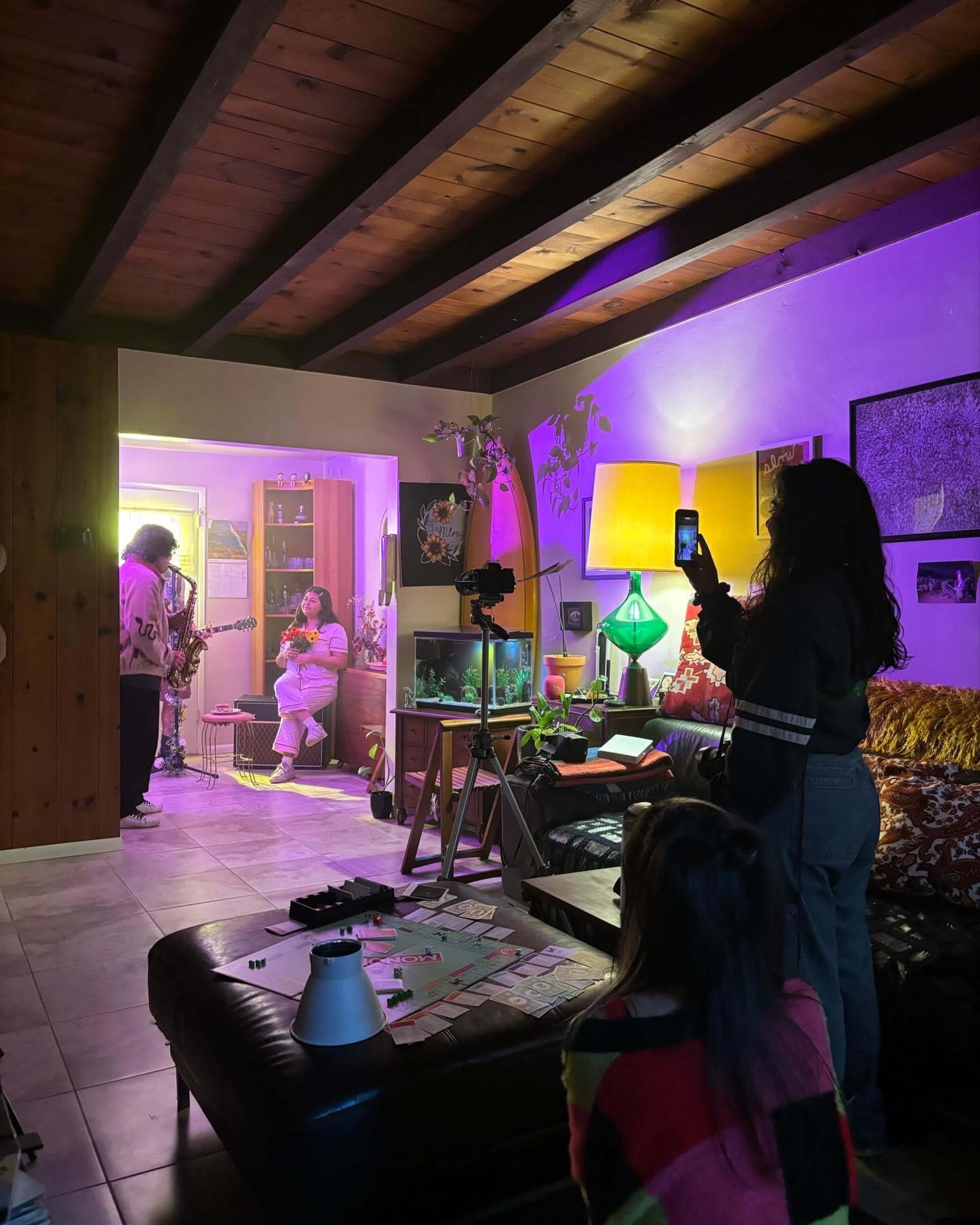 A group of people gathers in a cozy living room with colorful lighting, as one person plays an instrument while another records the scene with a phone.