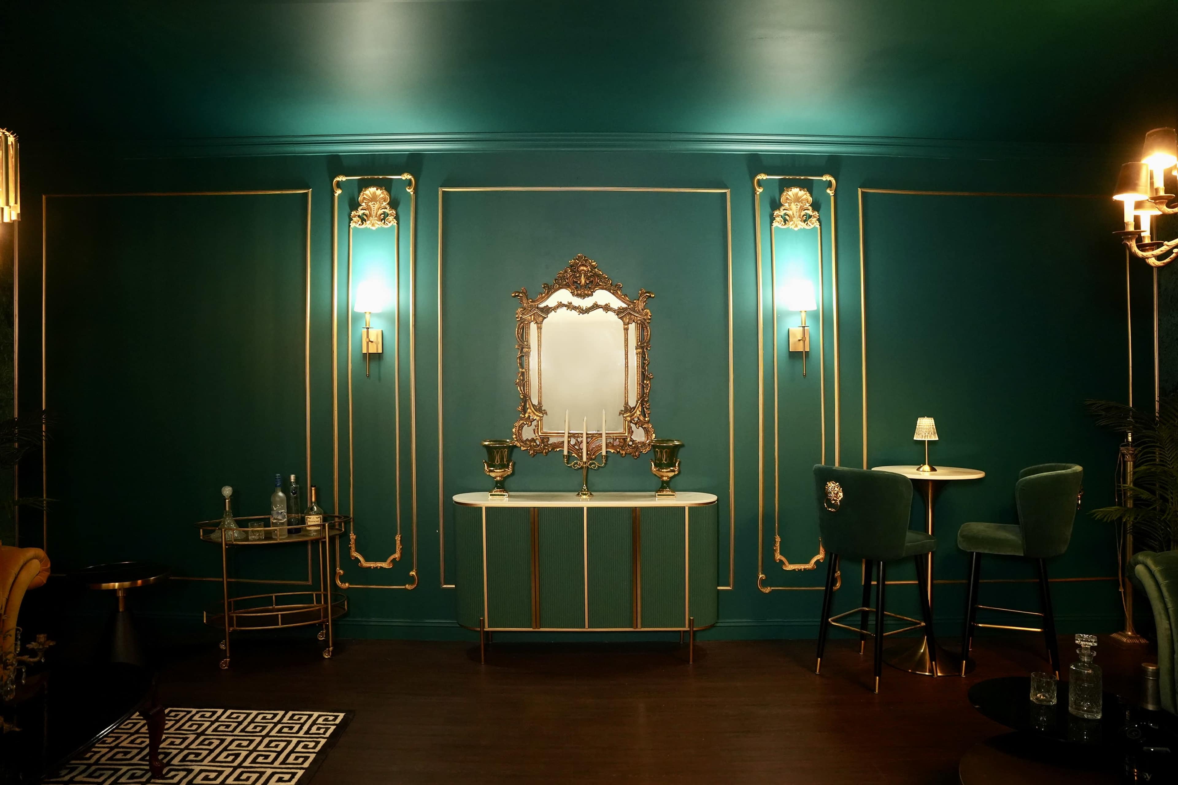A luxurious room featuring a teal-colored wall with gold trim, a large ornate mirror, a decorative table, and elegant seating.