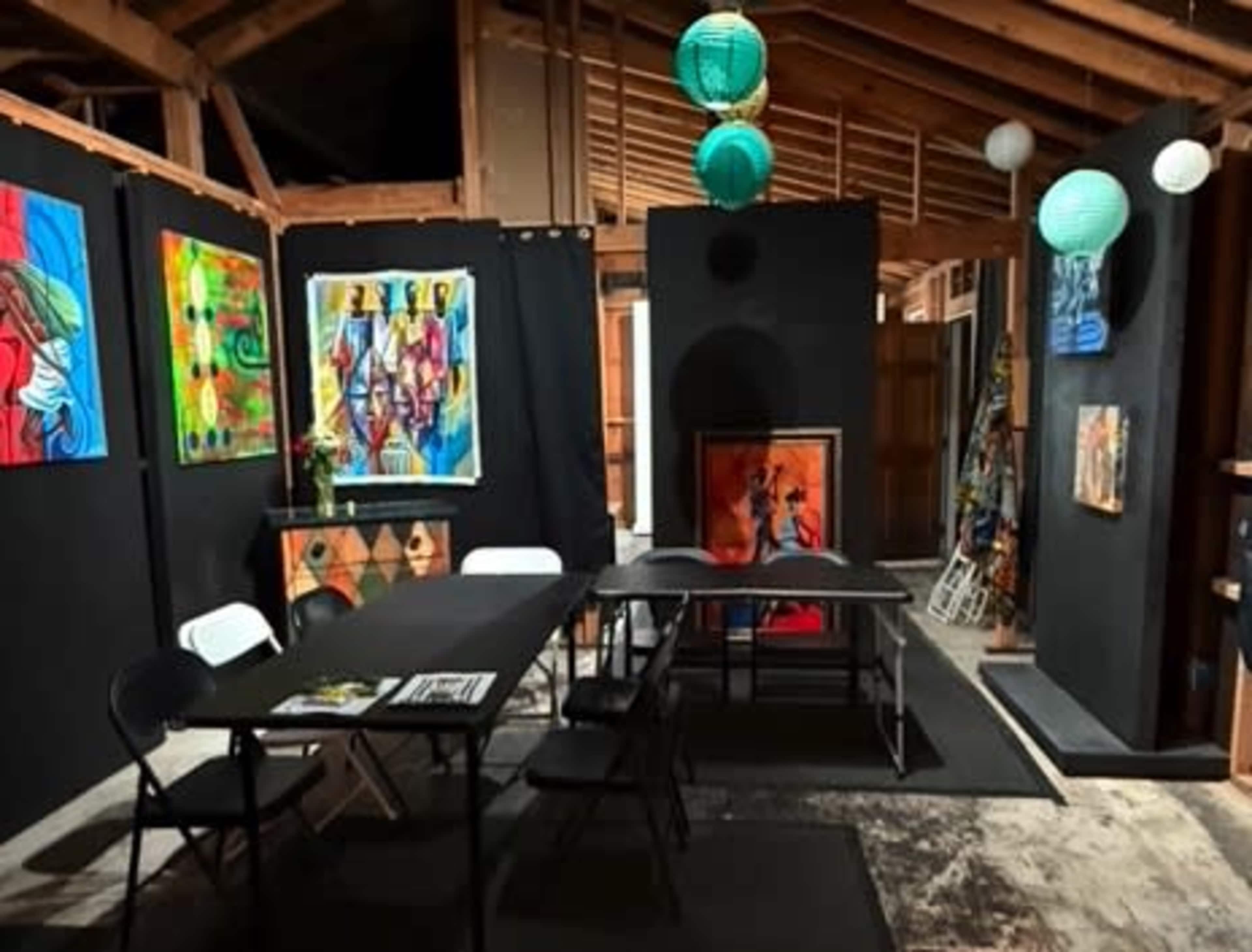 The image depicts an art studio with colorful paintings displayed on black walls and a table with chairs set up in the middle of the room.