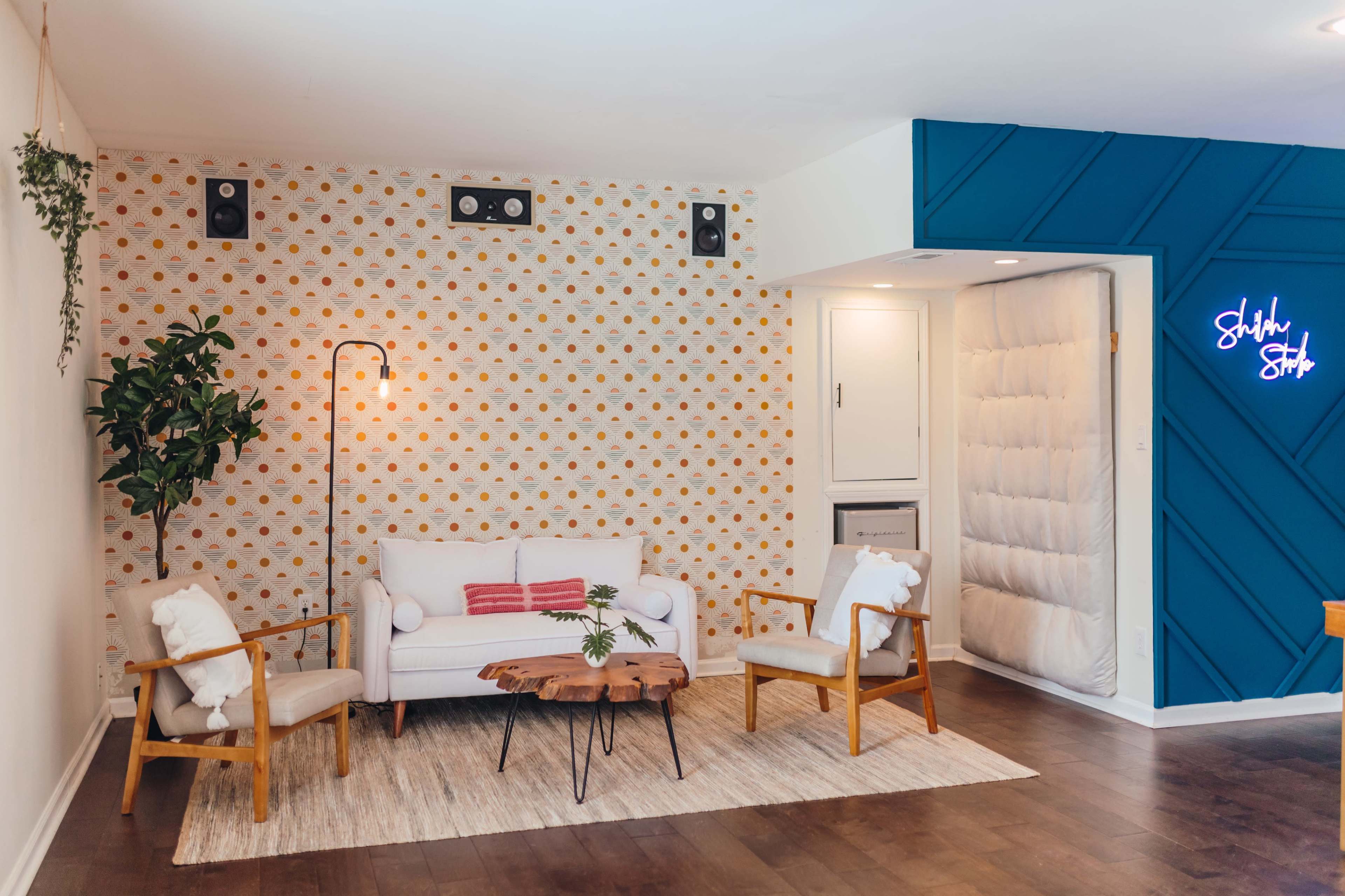 The image shows a modern living room with a patterned wallpaper, a white sofa, two wooden chairs with cushions, a round wooden coffee table, and a potted plant beside a floor lamp.