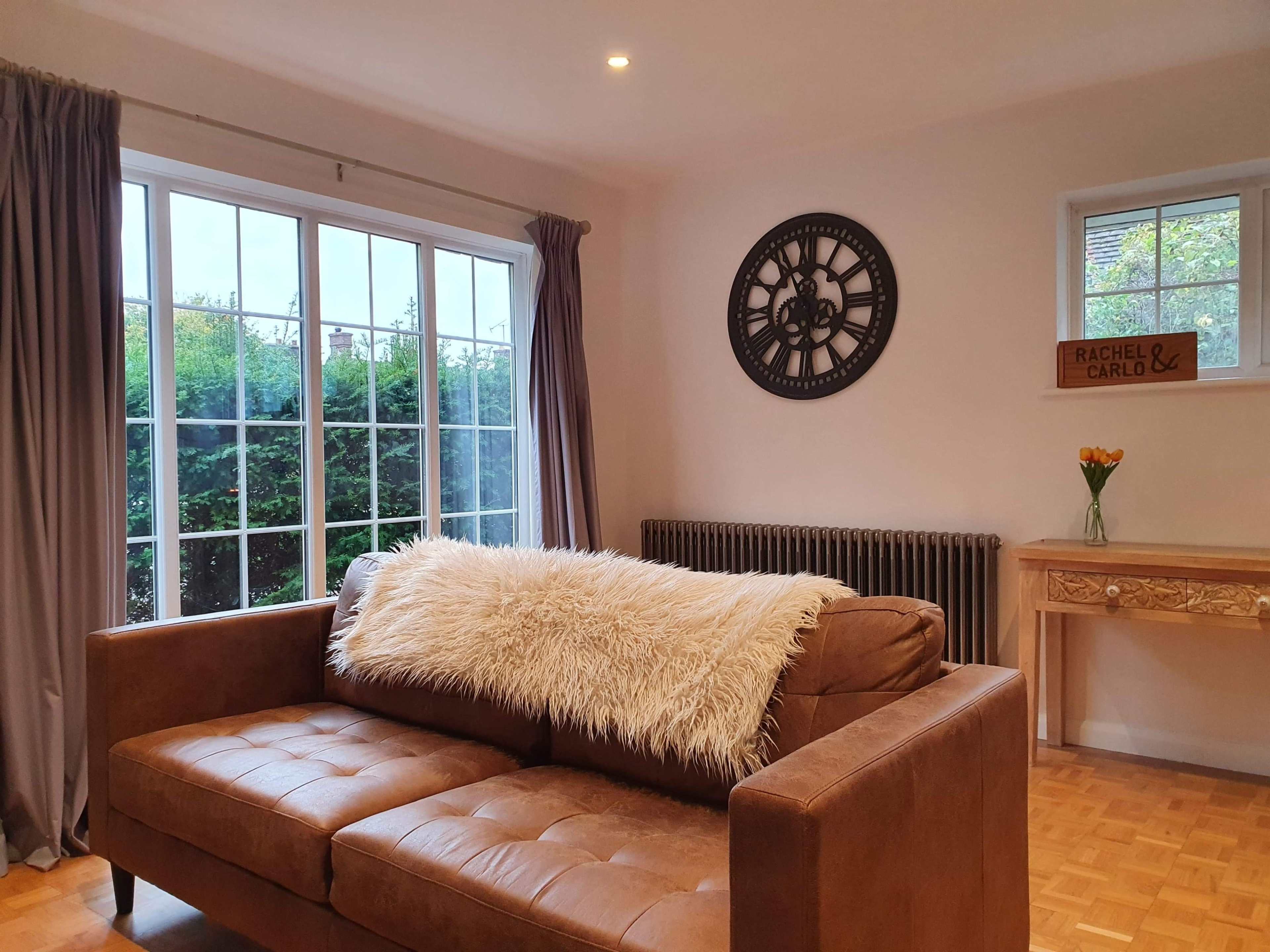 A living room with a brown leather sofa, a decorative wall with a clock, and a window partially covered by curtains, alongside a wooden table with a vase of flowers.