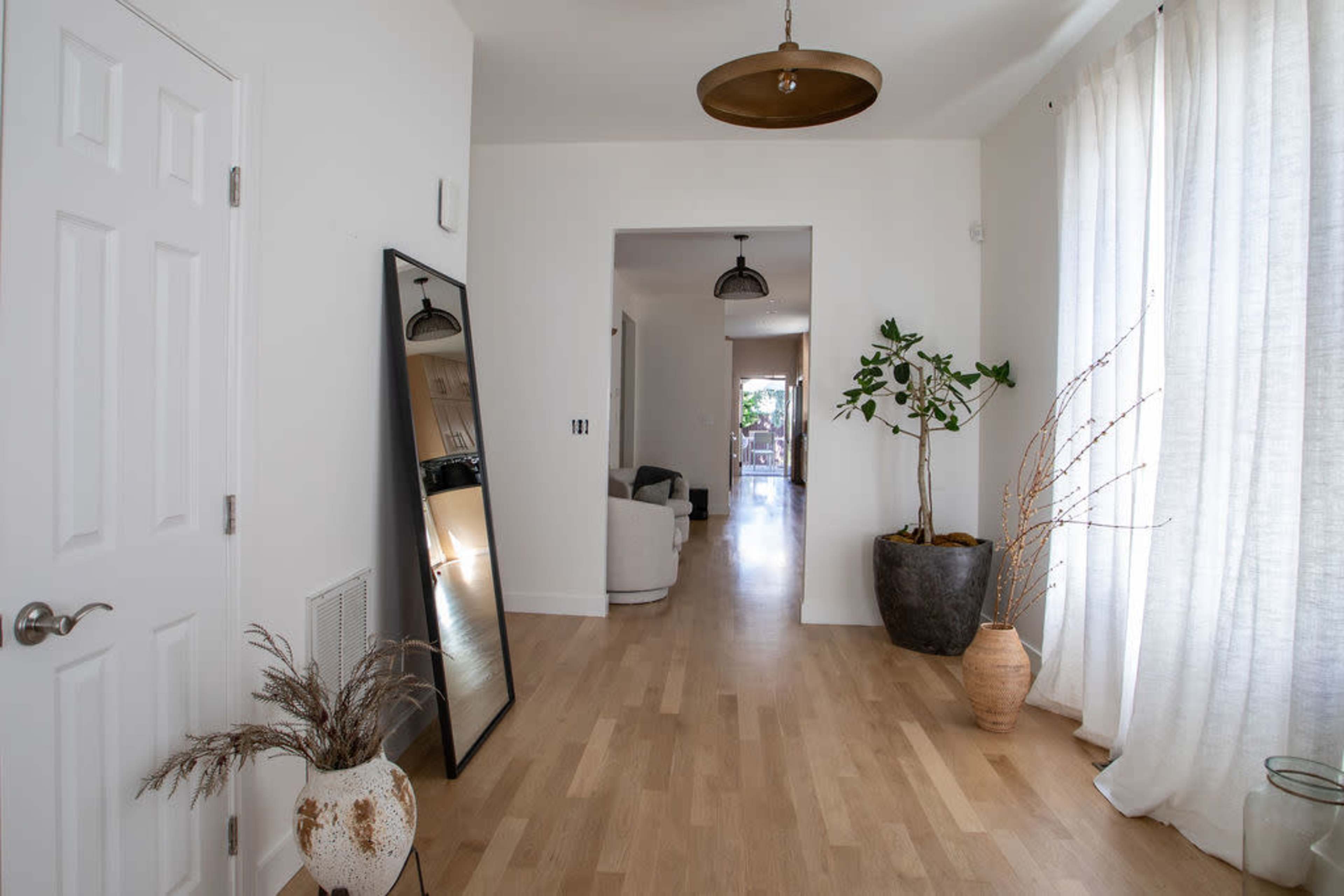 A bright hallway with a large mirror on the left, potted plants, and wood flooring leading to a living area in the background.