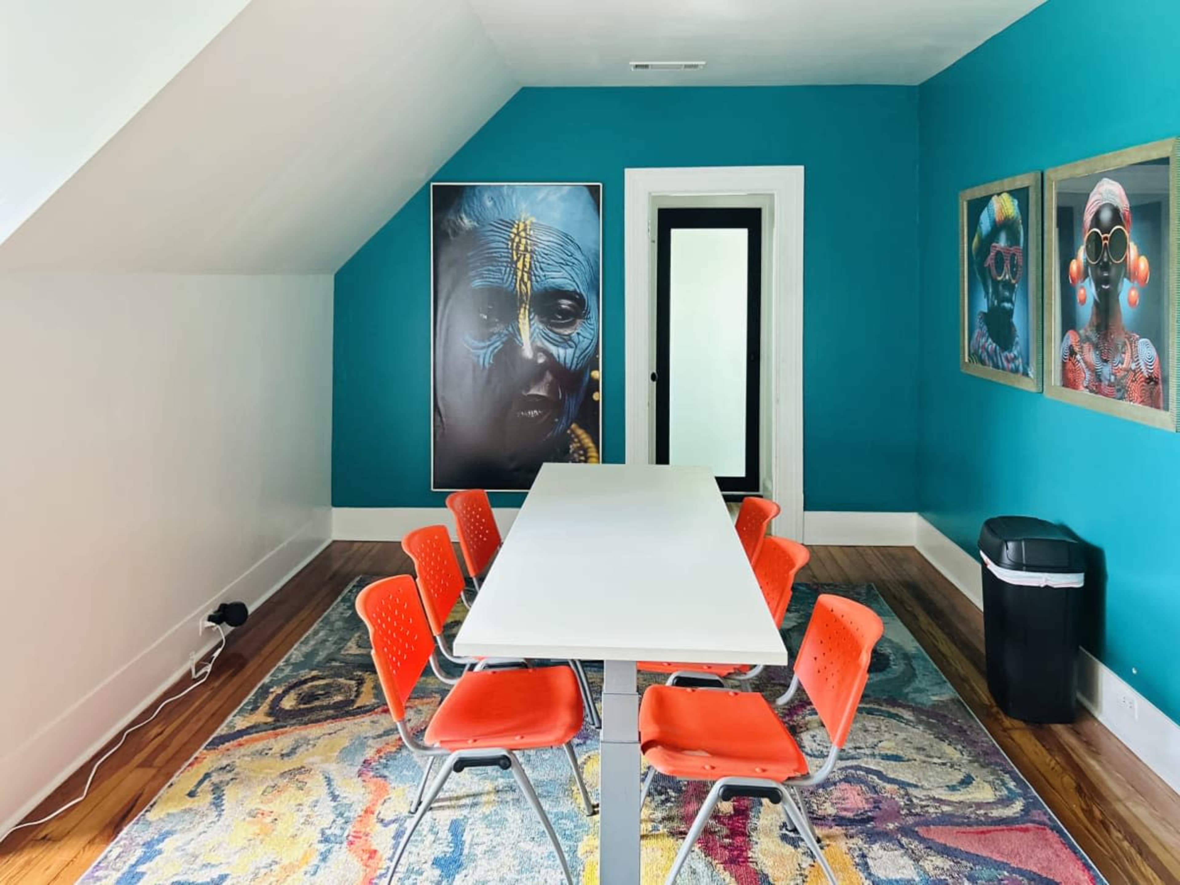 A bright room features a white table surrounded by orange chairs, with colorful artwork on the walls and a black trash can in the corner.