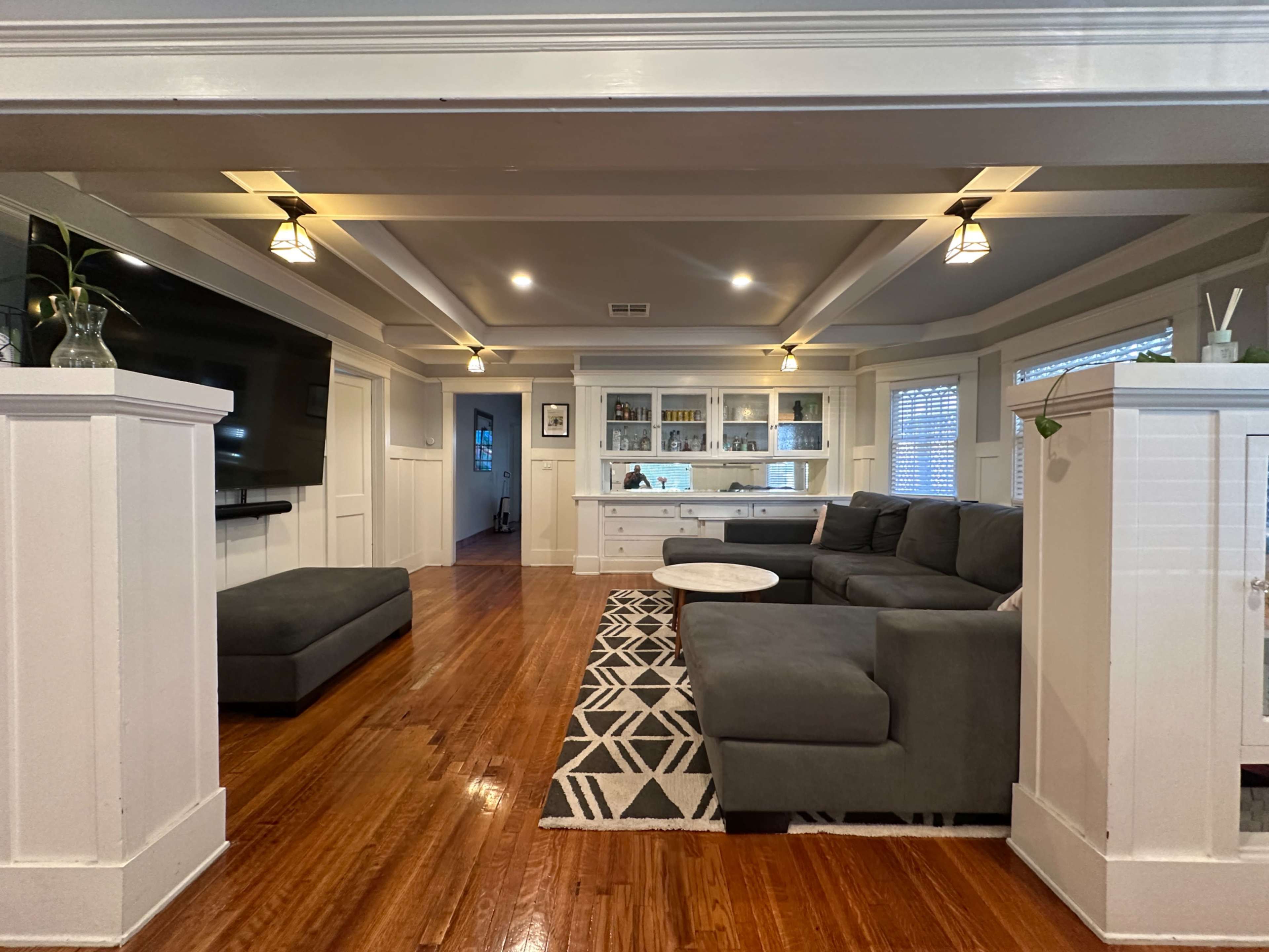 The image shows a modern living room with a large sectional sofa, a light rug, and a built-in shelving unit in the background.