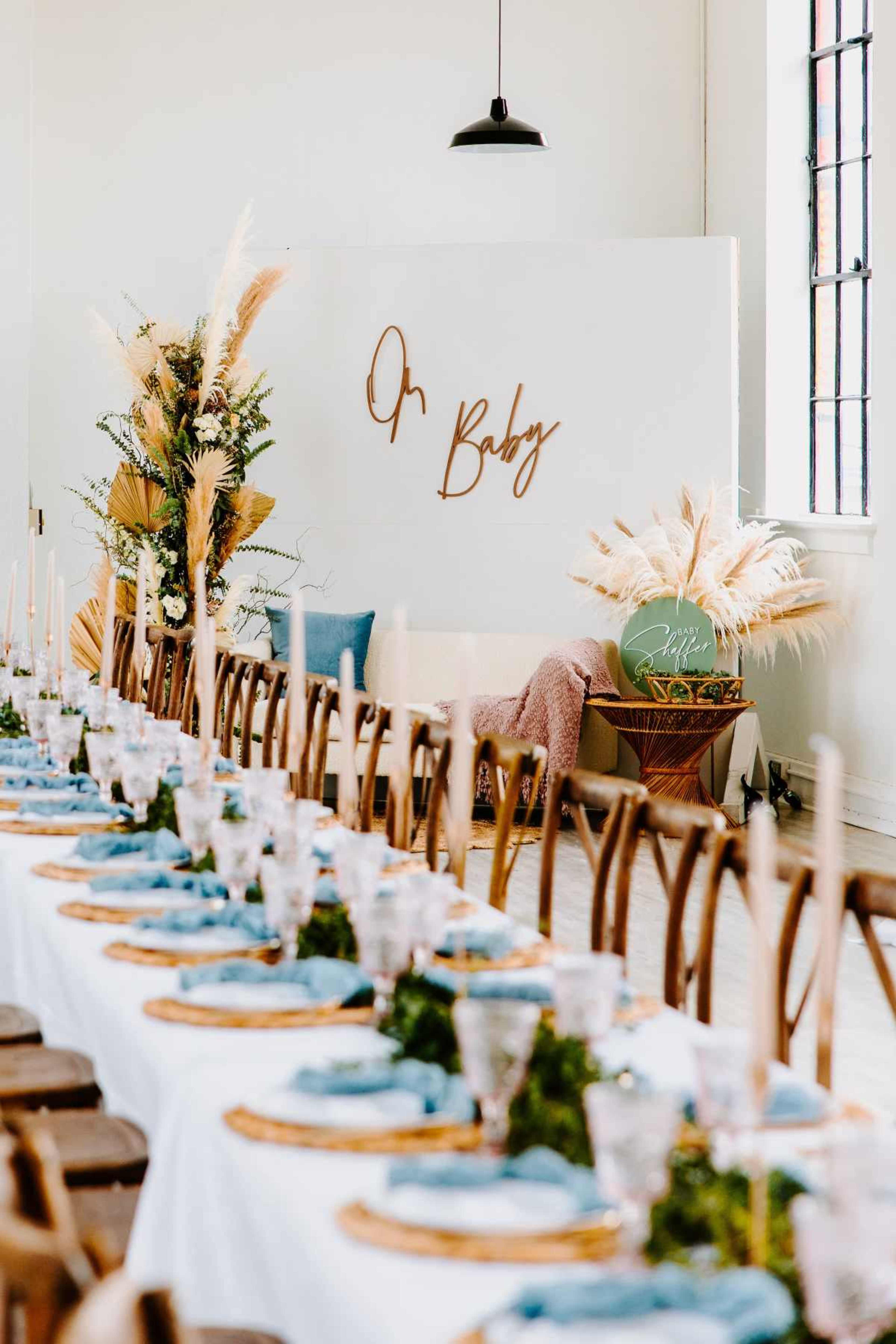 A long dining table is set with blue plates and glassware in a room featuring a decorated wall with the words "Oh Baby."