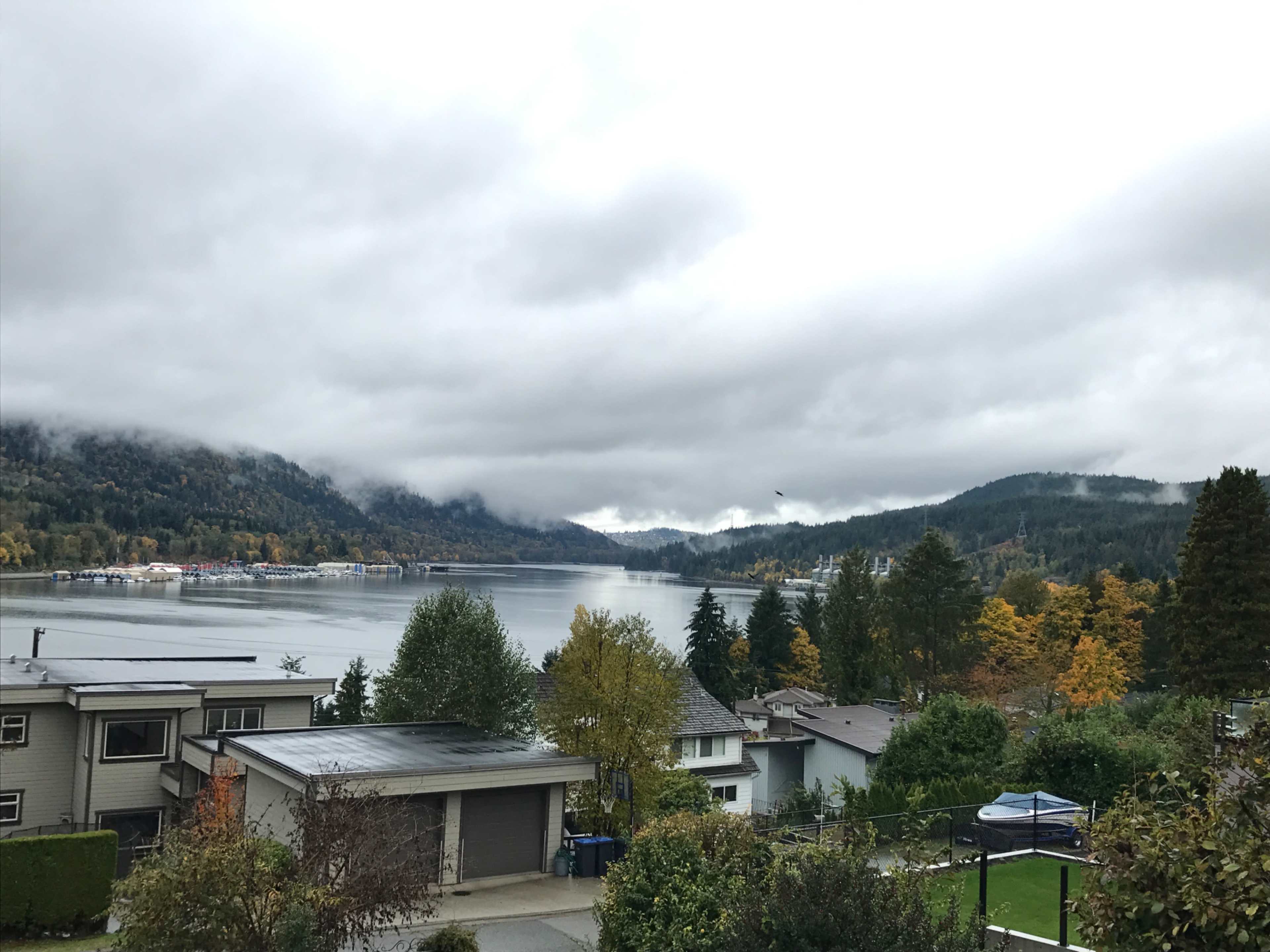 A body of water is surrounded by hills and houses under a cloudy sky with patches of autumn foliage.