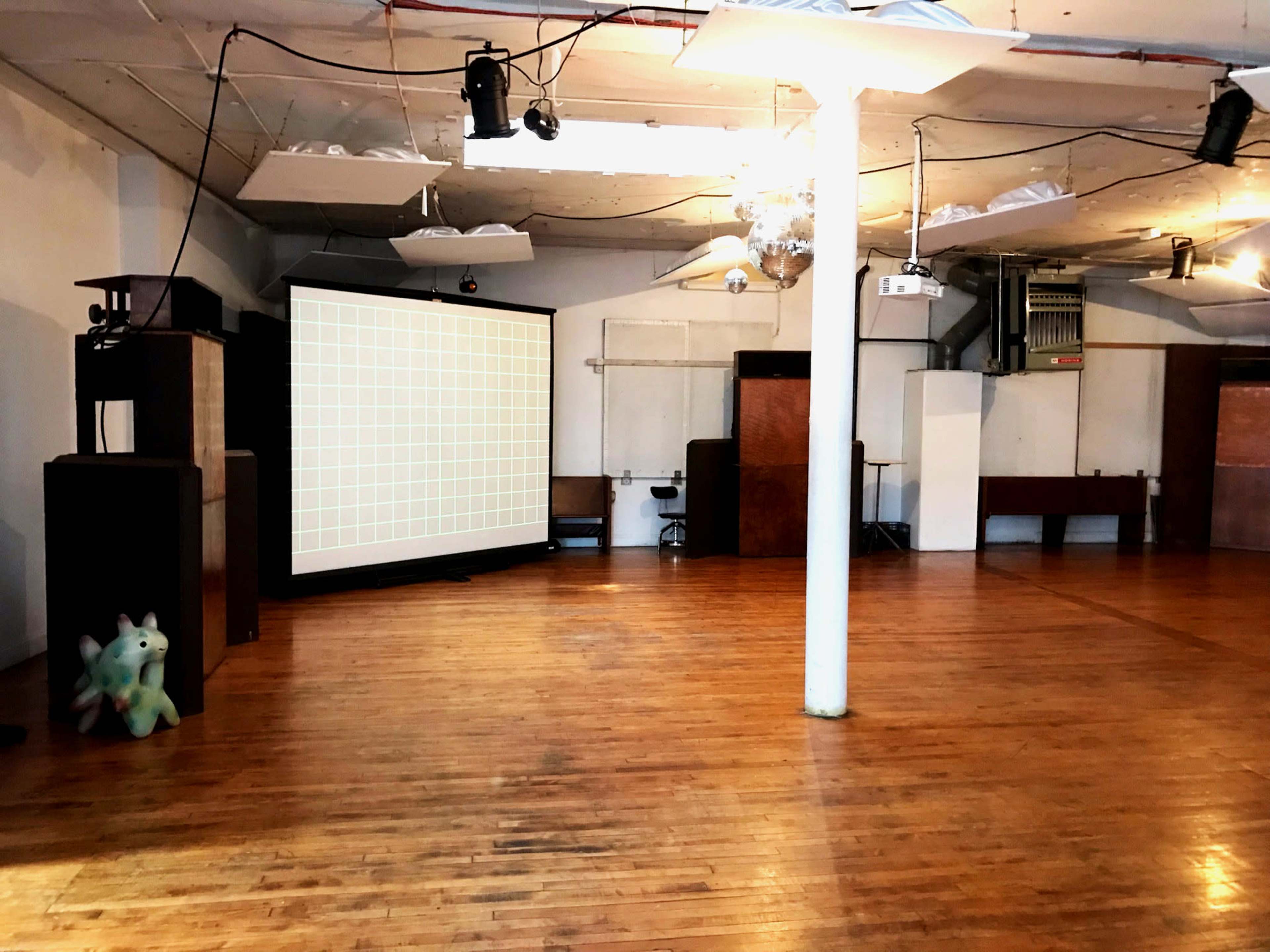 An empty room with hardwood floors features a large projection screen, a white column, and various wooden furniture pieces against the walls.