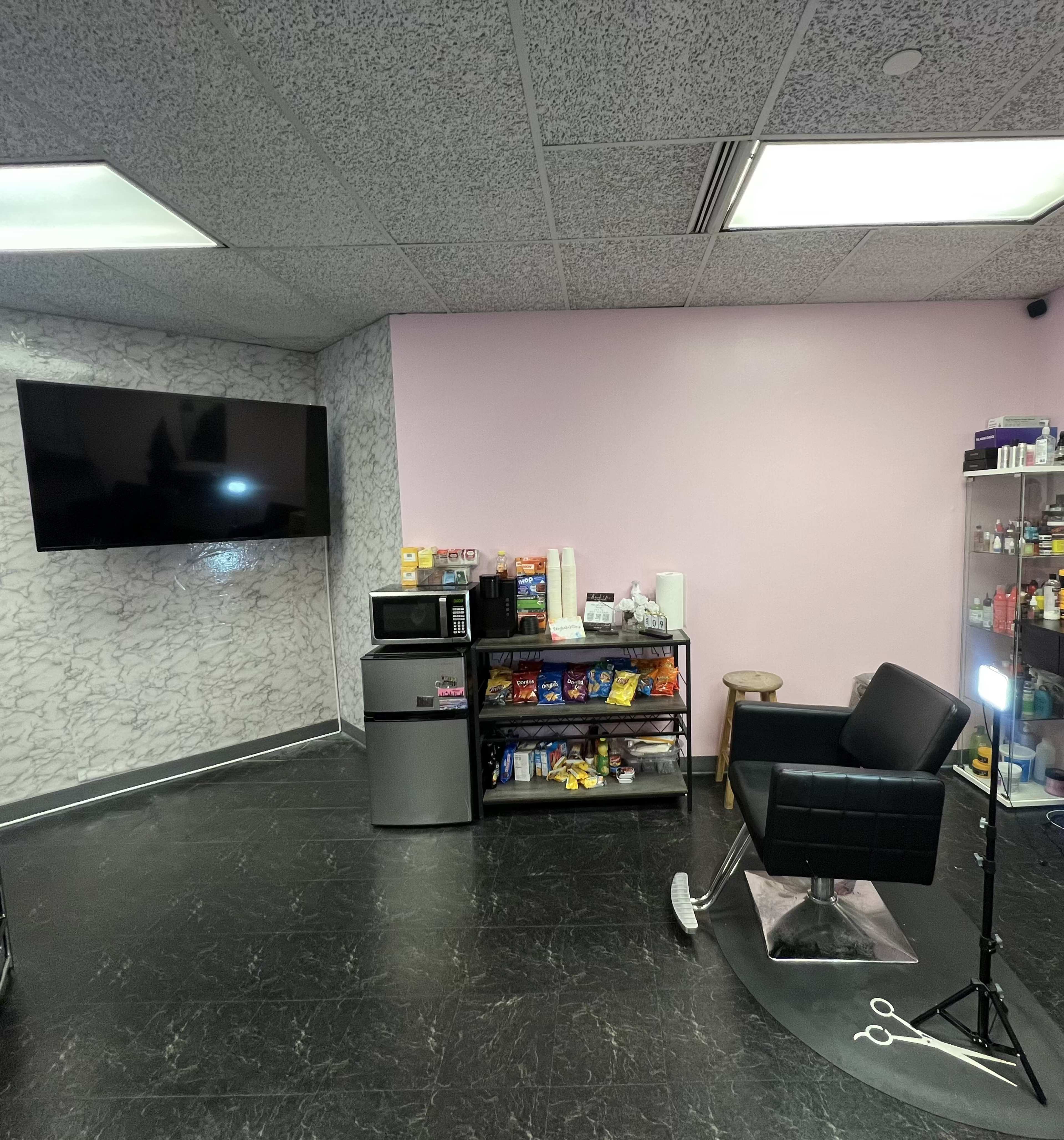 A salon interior with a black barber chair, a television on the wall, a display shelf filled with products, and a small kitchenette area with a microwave and snacks.