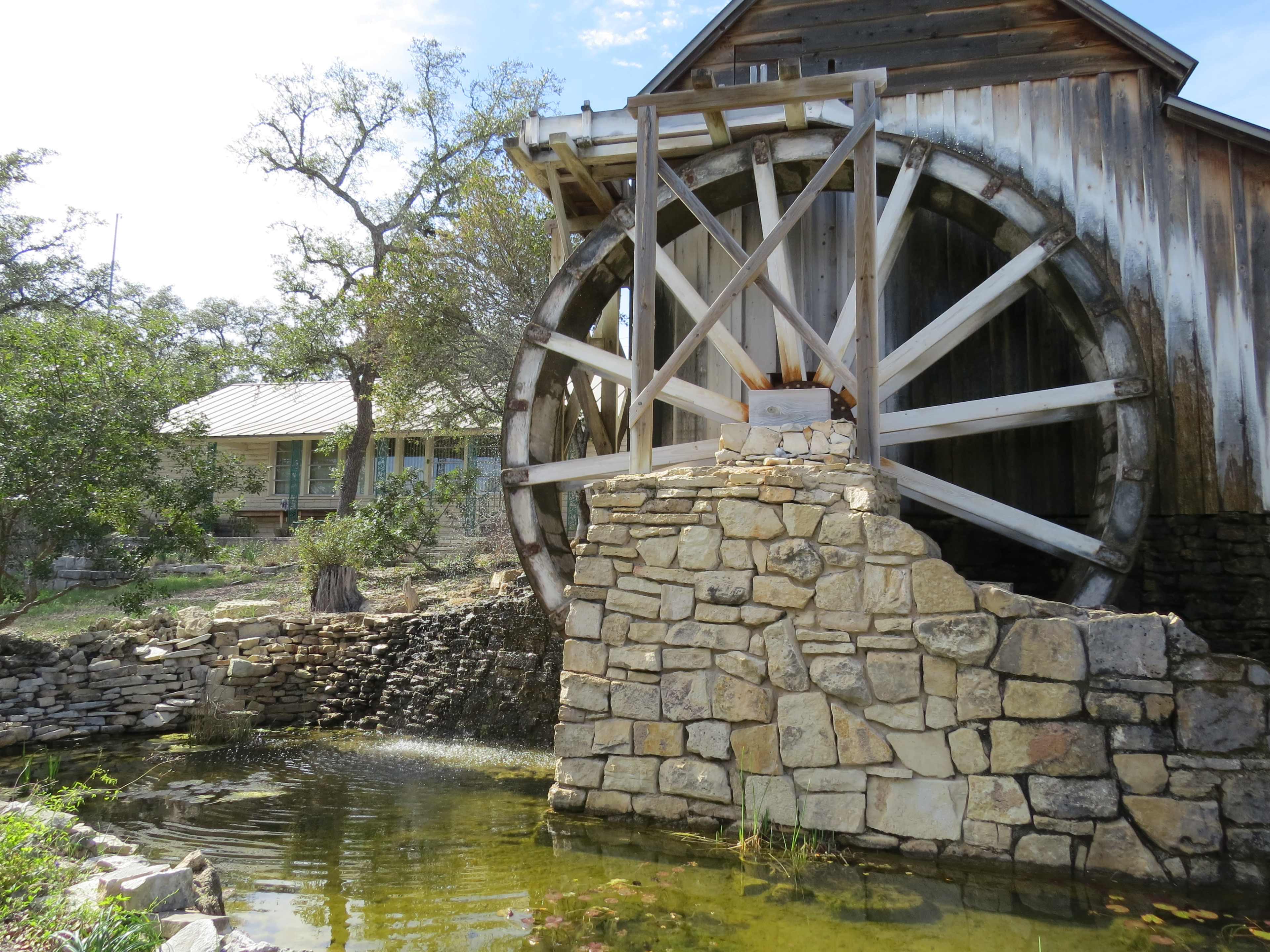 A wooden waterwheel is mounted on a stone structure beside a small pond and greenery.