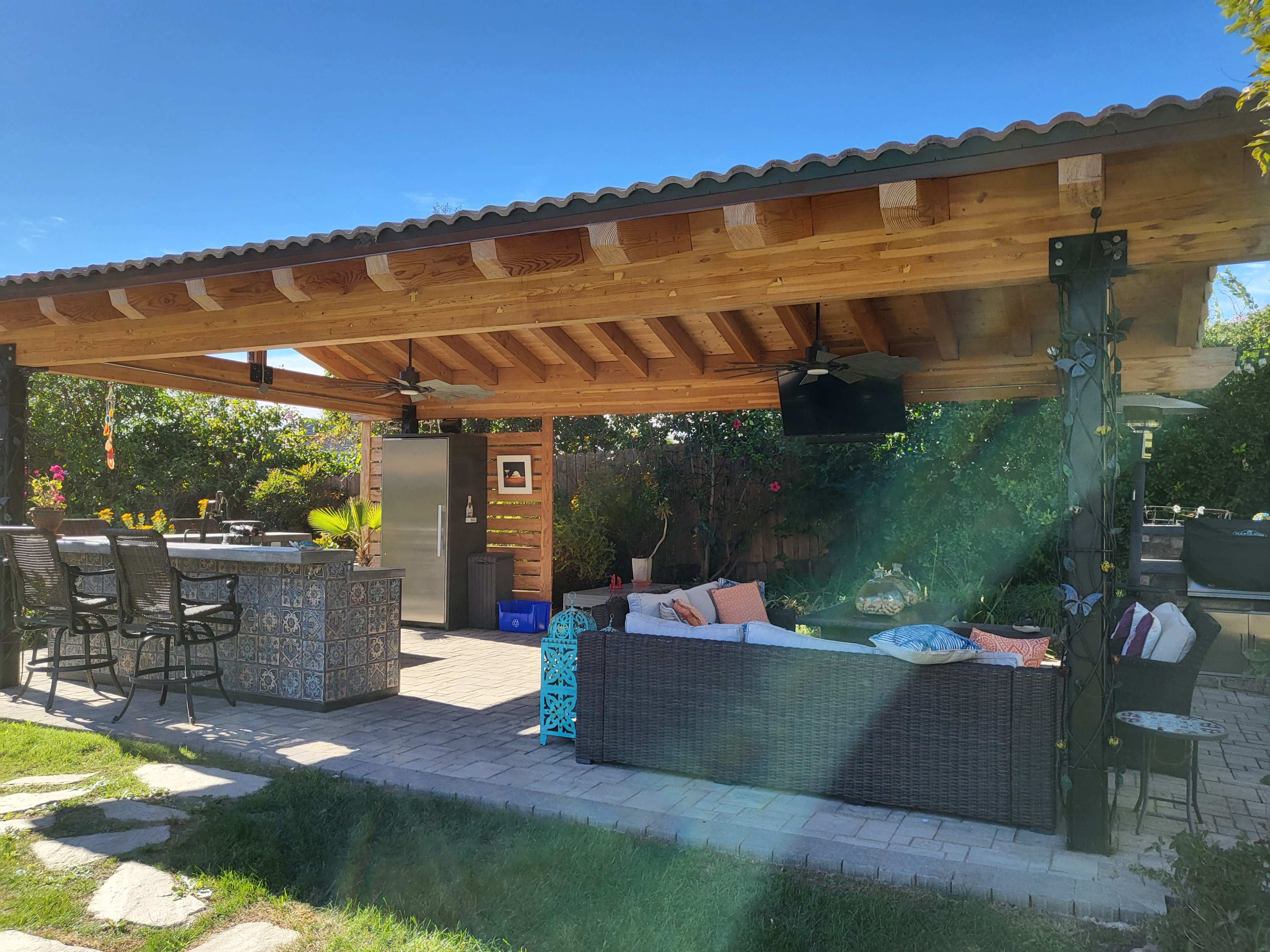 The image shows an outdoor patio area with a wooden roof, a bar with high stools, a large grill, and a cozy seating arrangement.