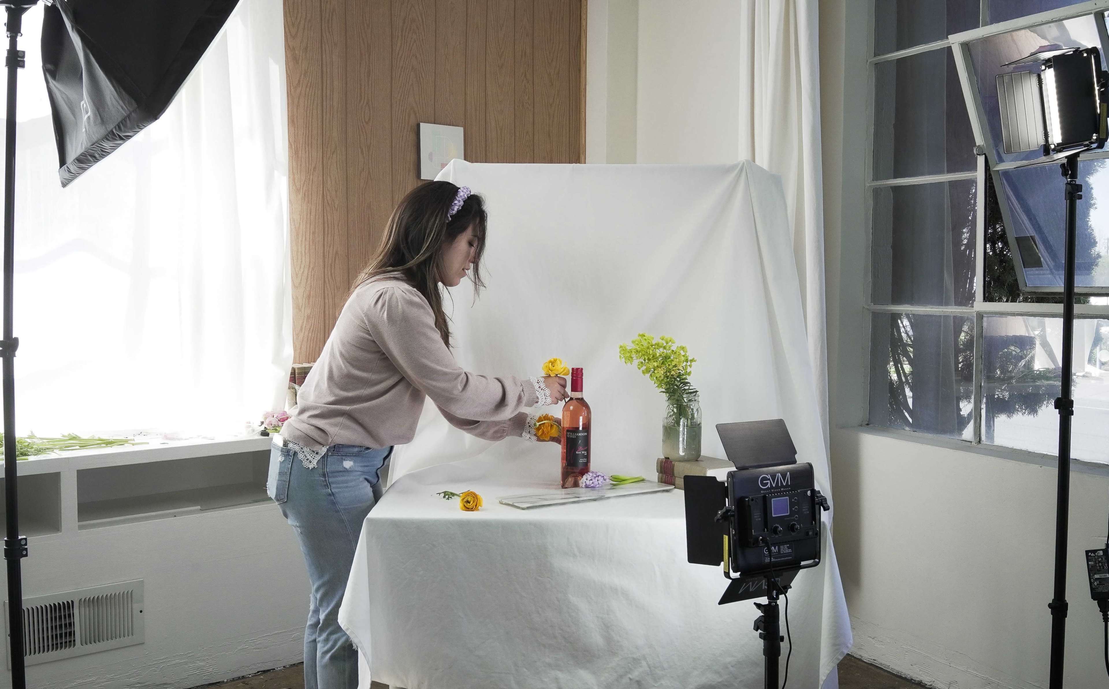 A woman is arranging flowers around a bottle on a table in a bright room set up for photography.