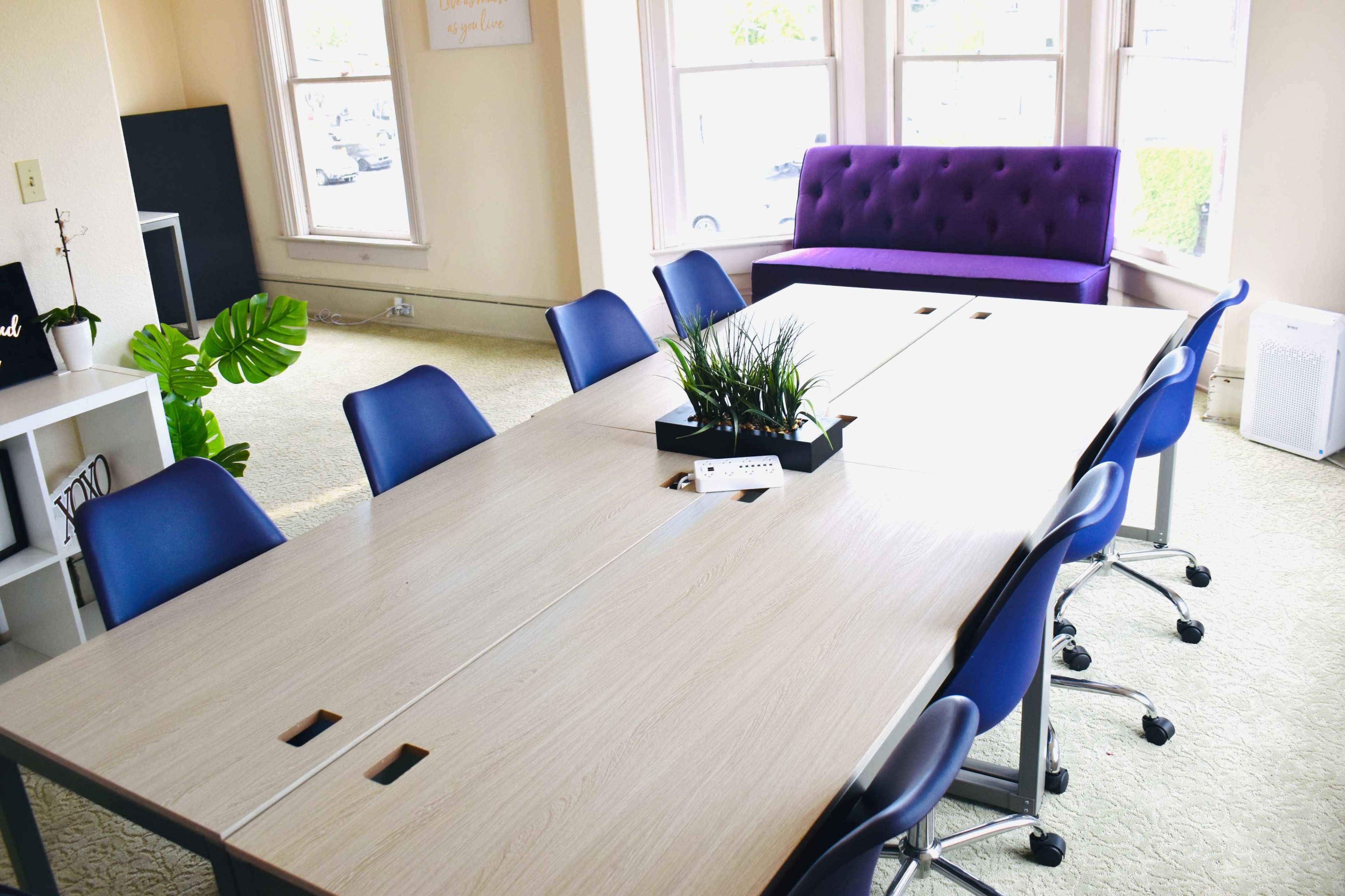 A large conference table with blue chairs is positioned in a bright room featuring a purple couch and a potted plant.