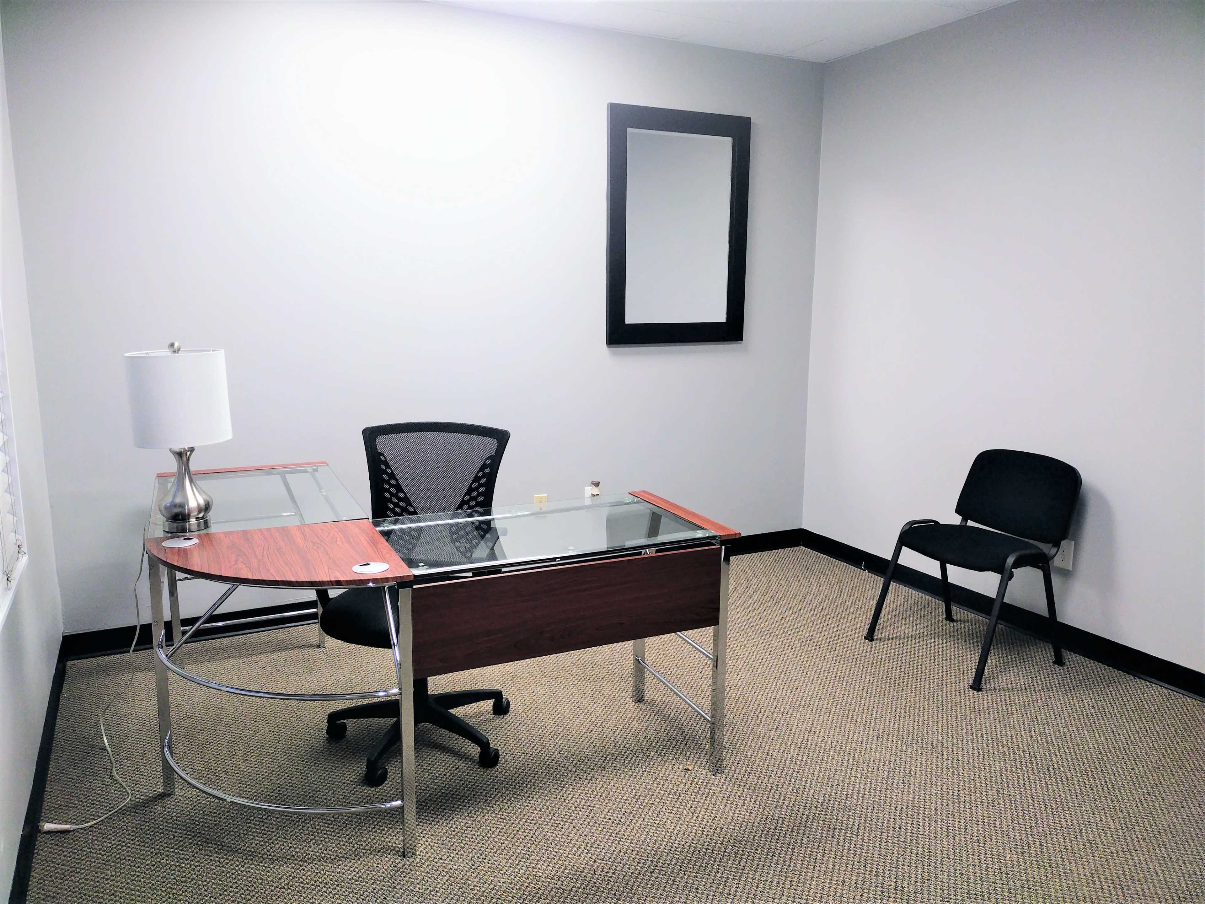 A sparsely furnished office with a glass-top desk, an office chair, a lamp, a mirror on the wall, and a single black chair.