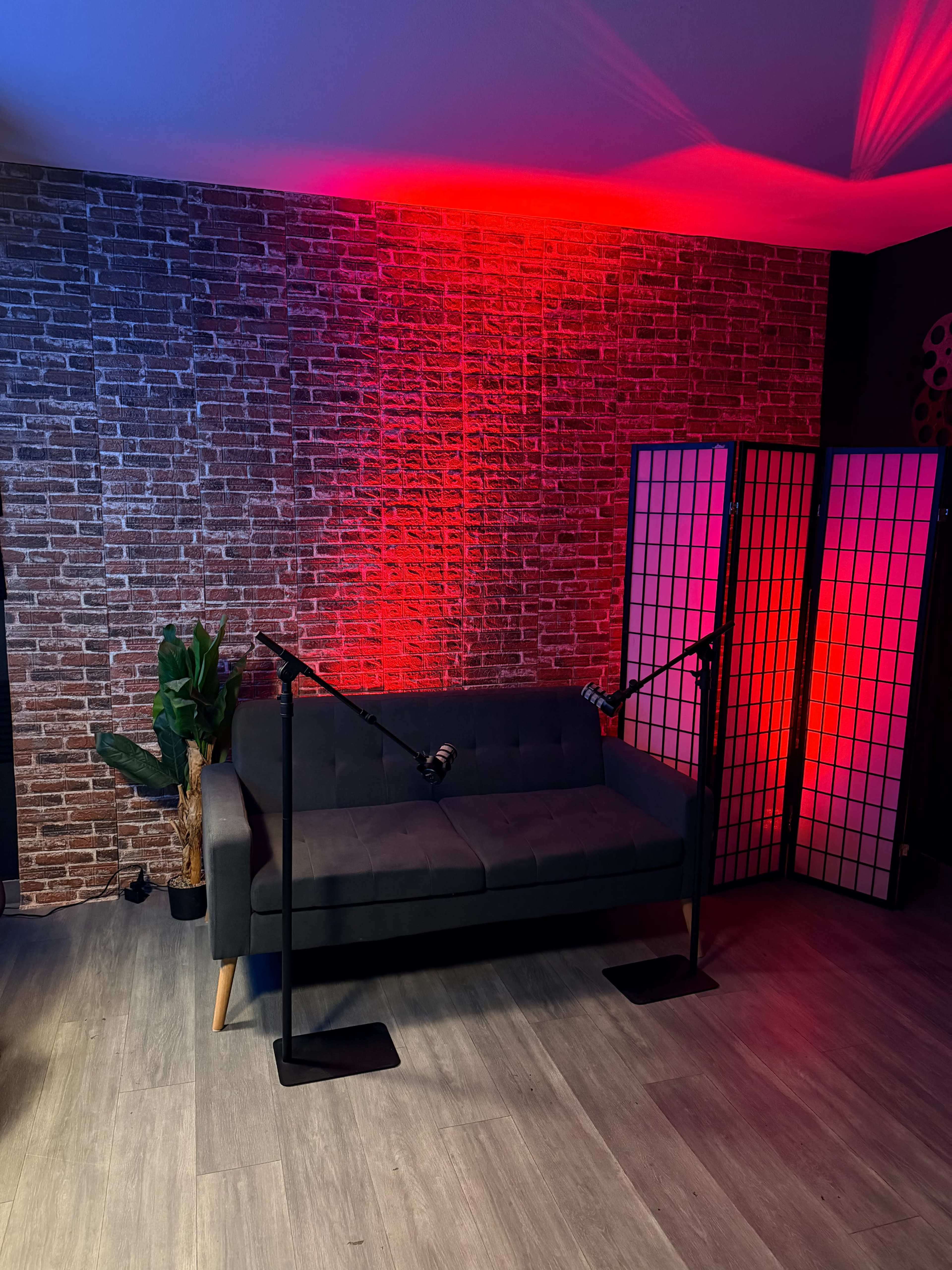 The scene features a dark gray couch positioned in front of a brick wall, with two black microphone stands beside it and colorful lighting creating a moody atmosphere.