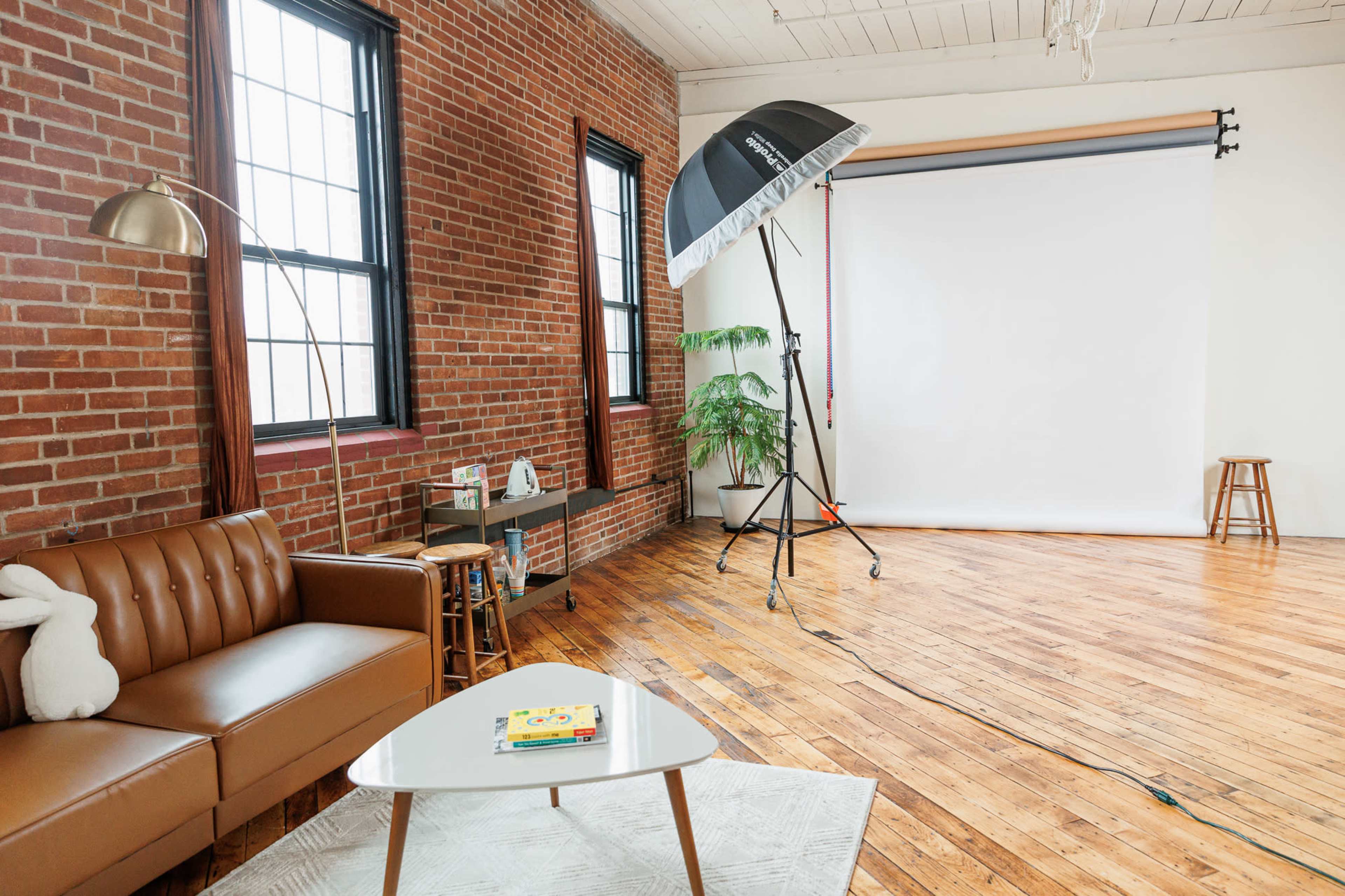 A spacious photography studio features a brown leather sofa, a bright white backdrop, and large windows with exposed brick walls.