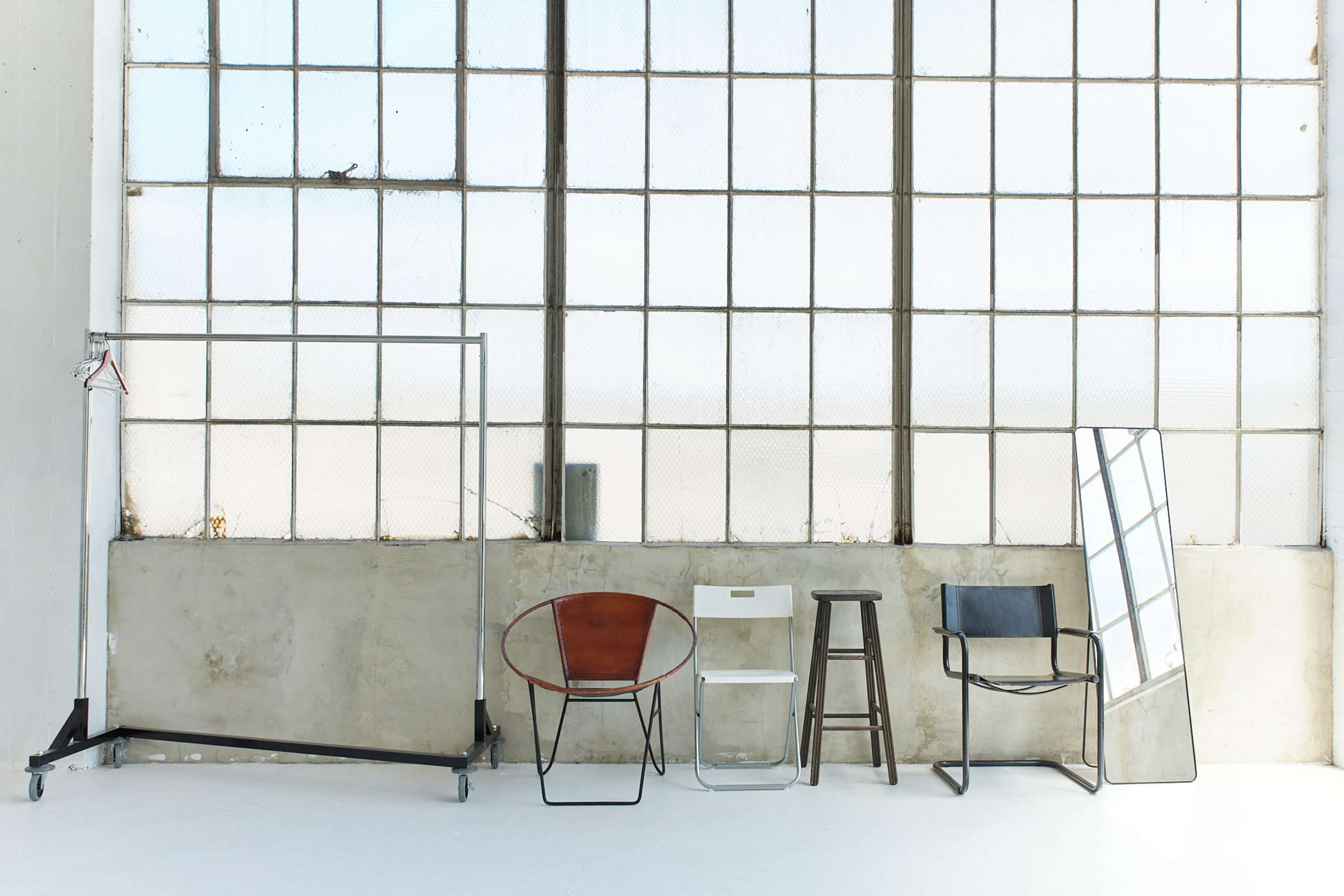 The image shows a row of five distinct chairs and a full-length mirror set against a backdrop of large industrial windows.