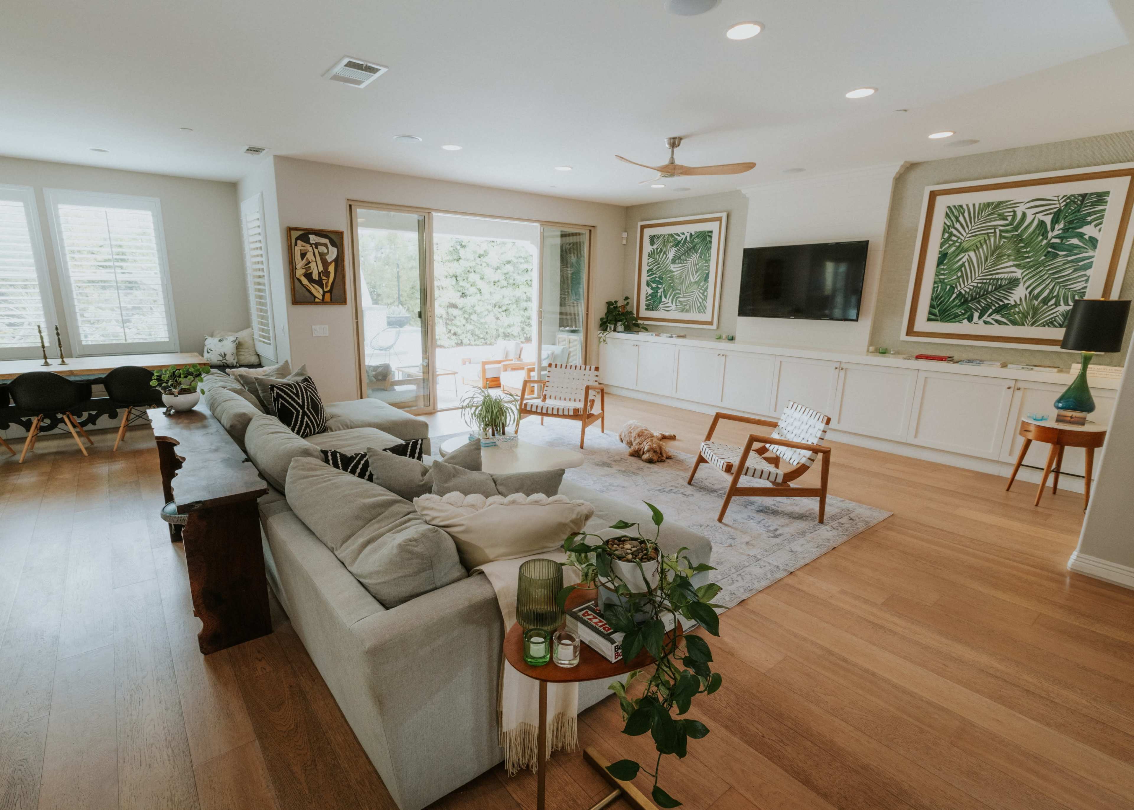 The image shows a spacious living room featuring a light-colored sofa, a coffee table with plants, and large windows letting in natural light.